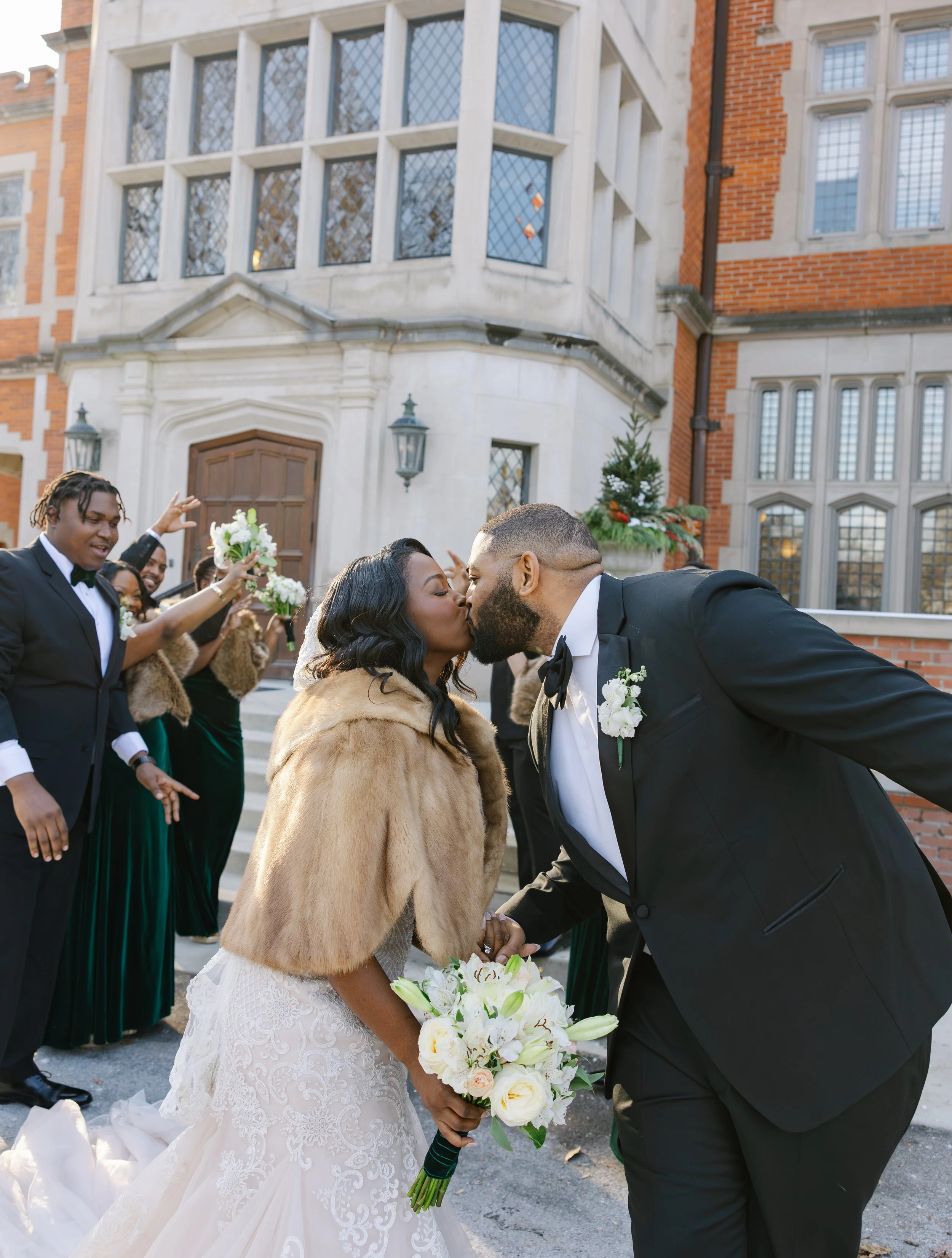 A bride and groom kiss during their wedding celebration outside a large, historic building, surrounded by friends and wedding party members cheering and holding flowers.