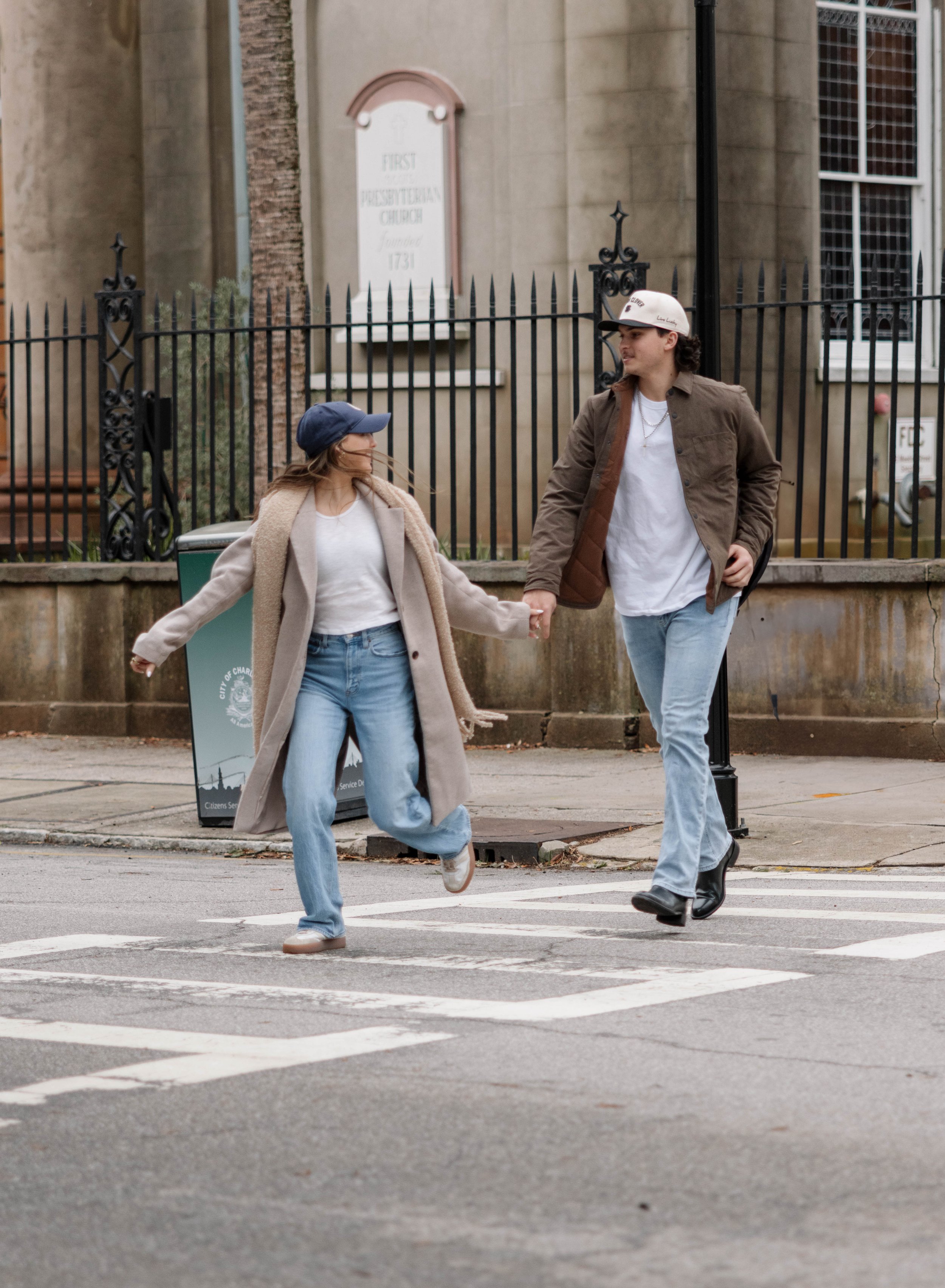 A young woman and a young man holding hands and crossing a street at a crosswalk, smiling and looking at each other, in front of a building with a black iron fence.