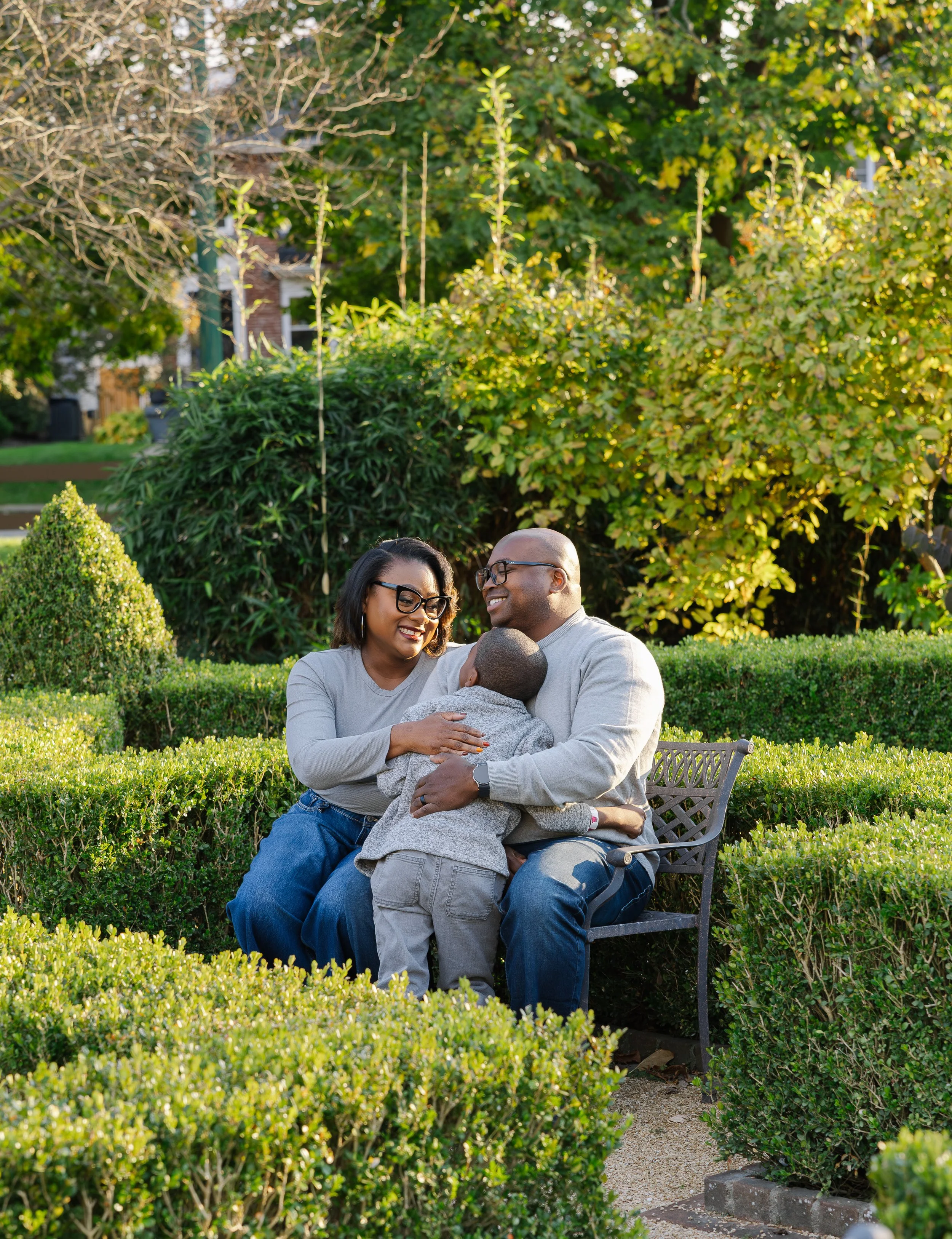 A happy African American family sitting on a bench in a park, embracing each other, surrounded by green bushes and trees with sunlight filtering through the foliage.