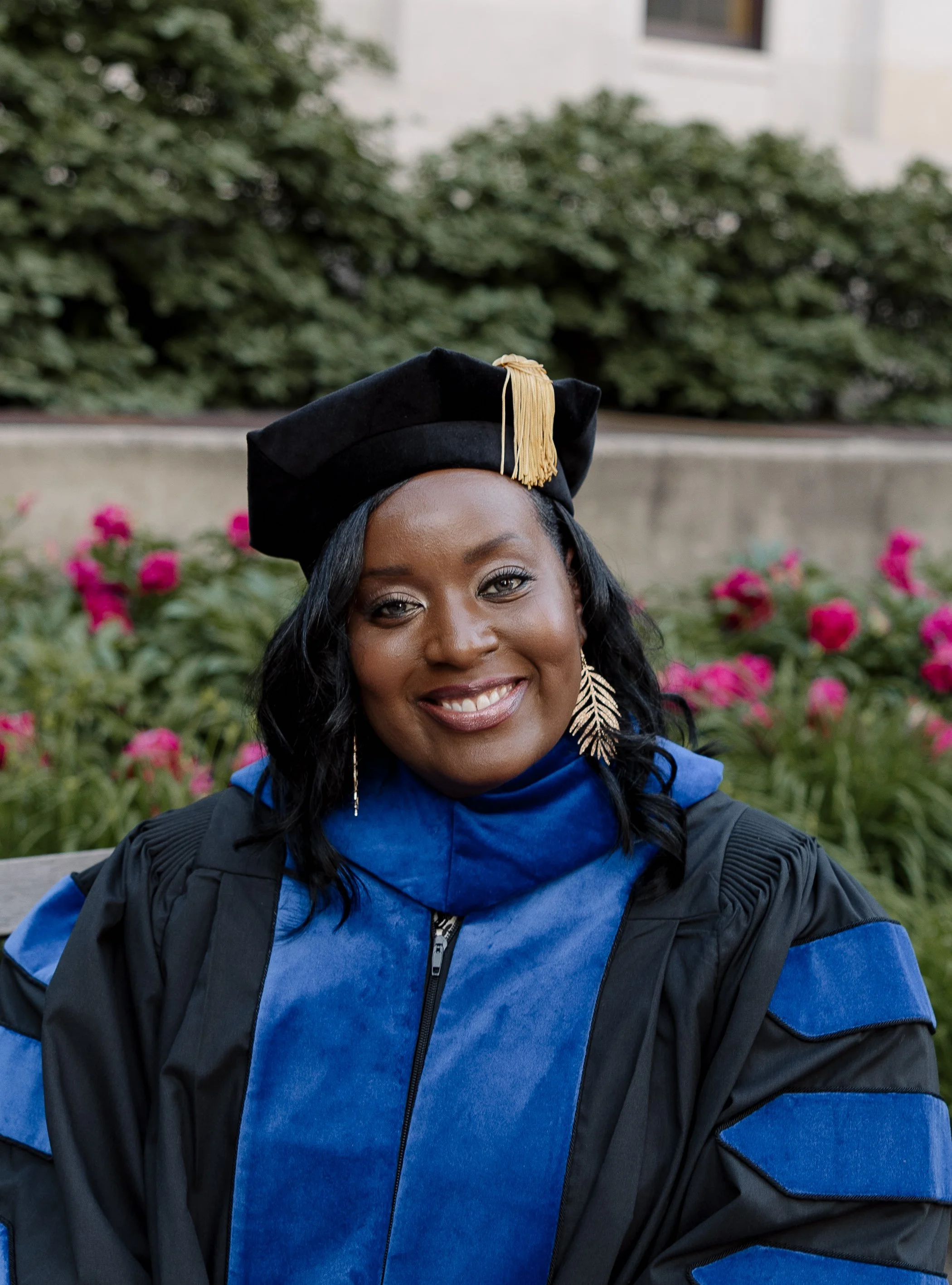Woman in academic regalia, wearing a black cap with a gold tassel and a blue and black gown, smiling outdoors with pink flowers in the background.
