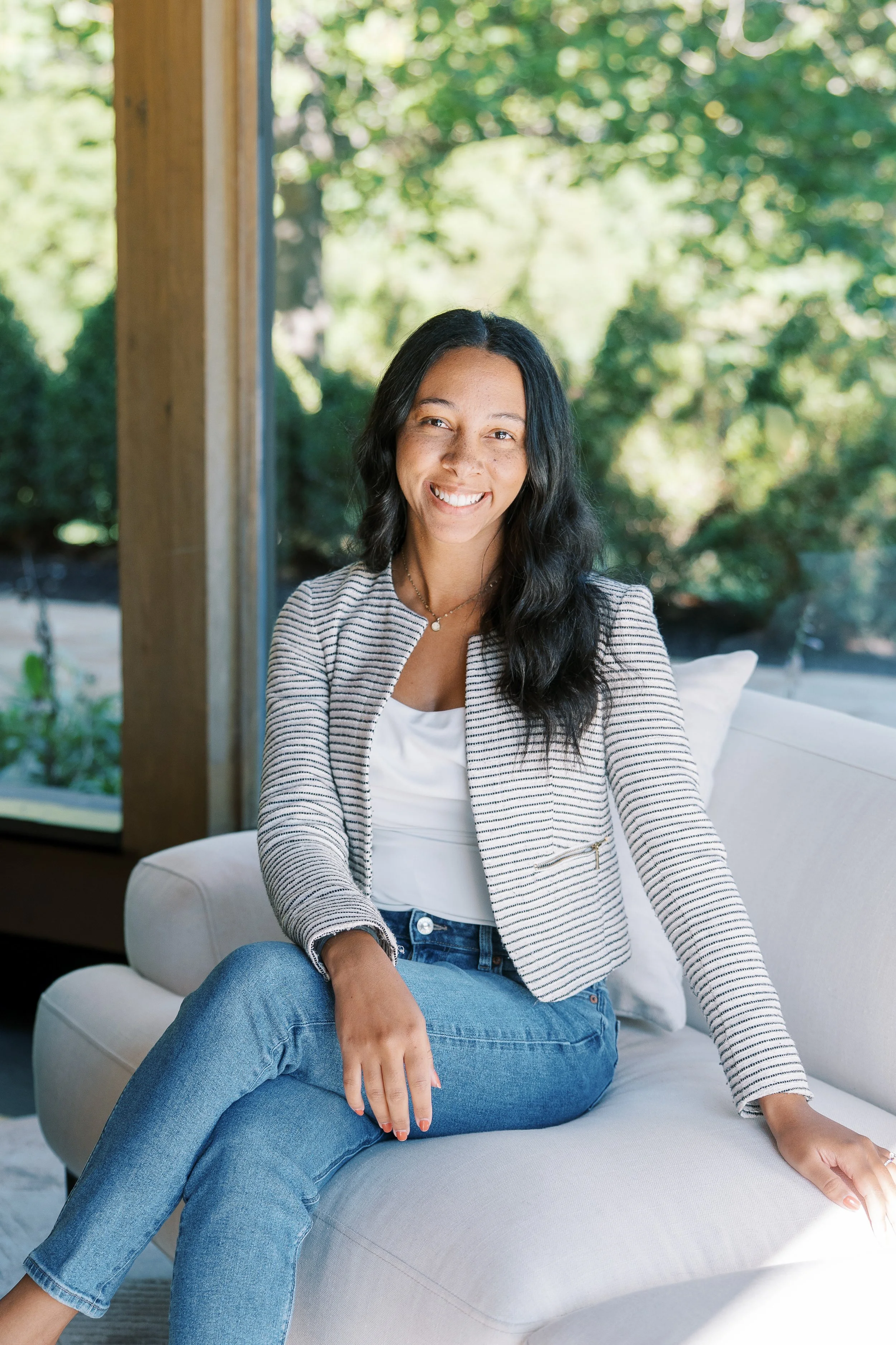 A woman with dark wavy hair smiling while sitting on a white sofa near large windows overlooking a green outdoor area.