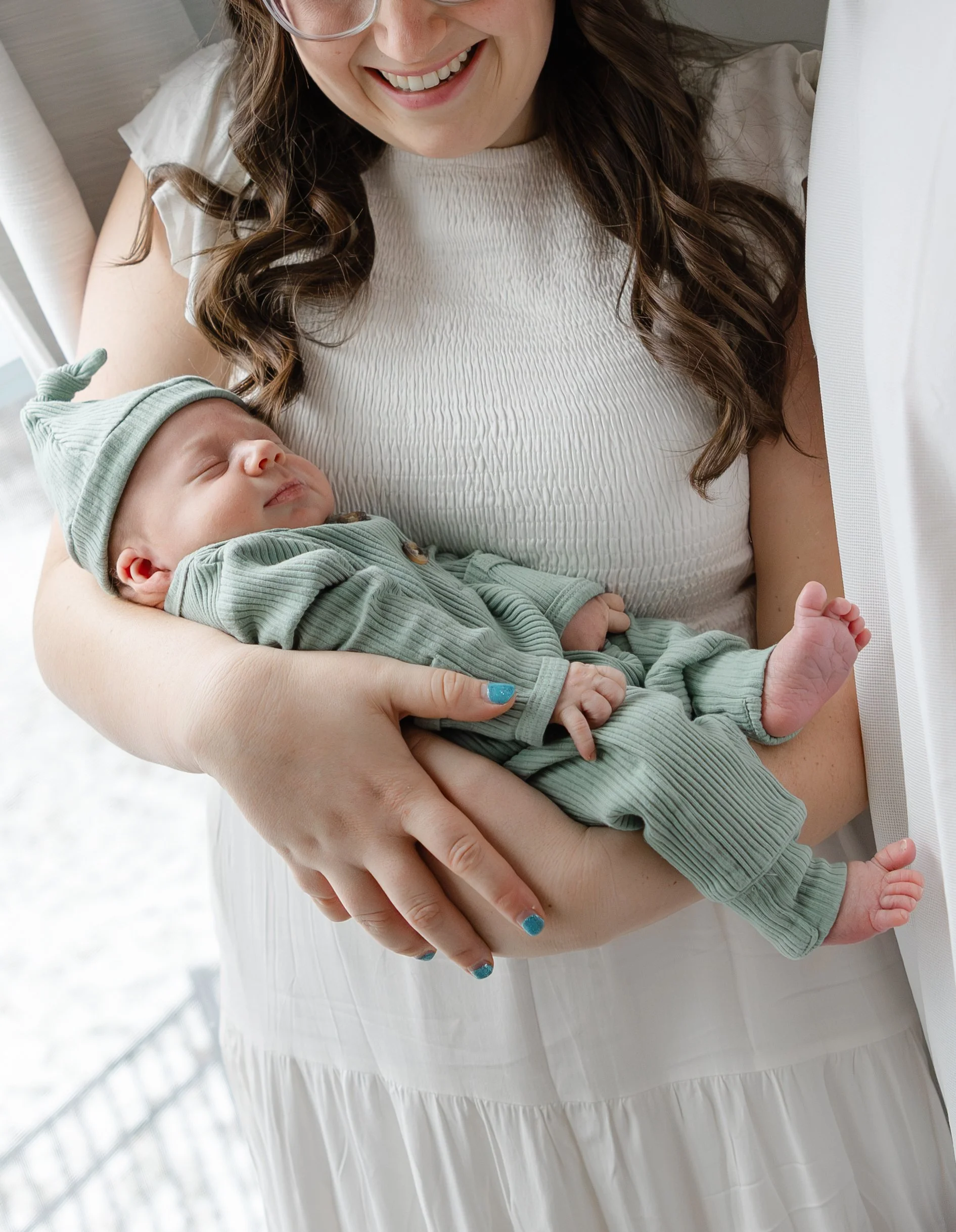 A woman happily holding a sleeping baby dressed in green with a matching green hat, indoors near a window with white curtains.
