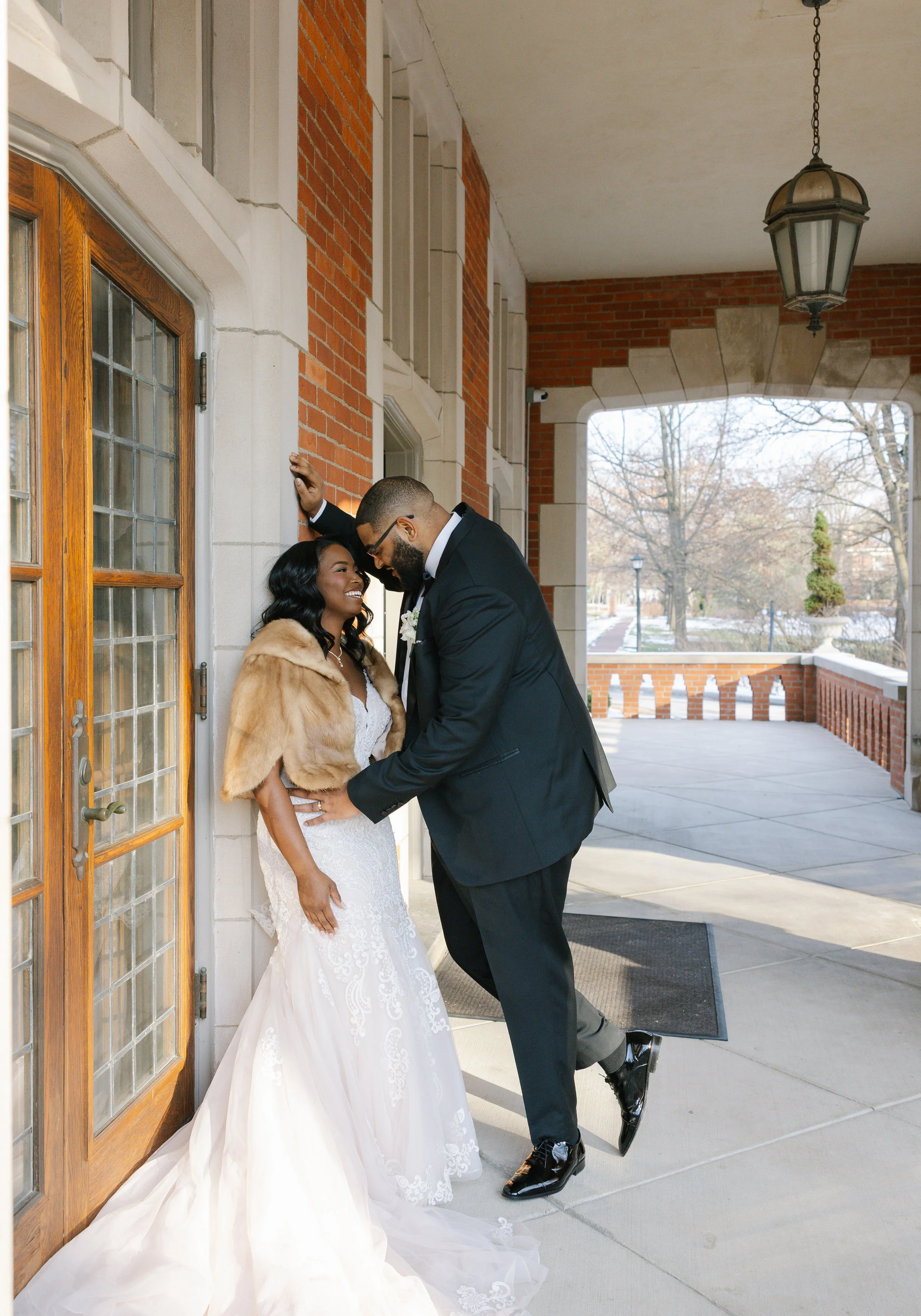 A newlywed couple smiling and embracing outside a brick building, with trees and a pathway visible in the background.