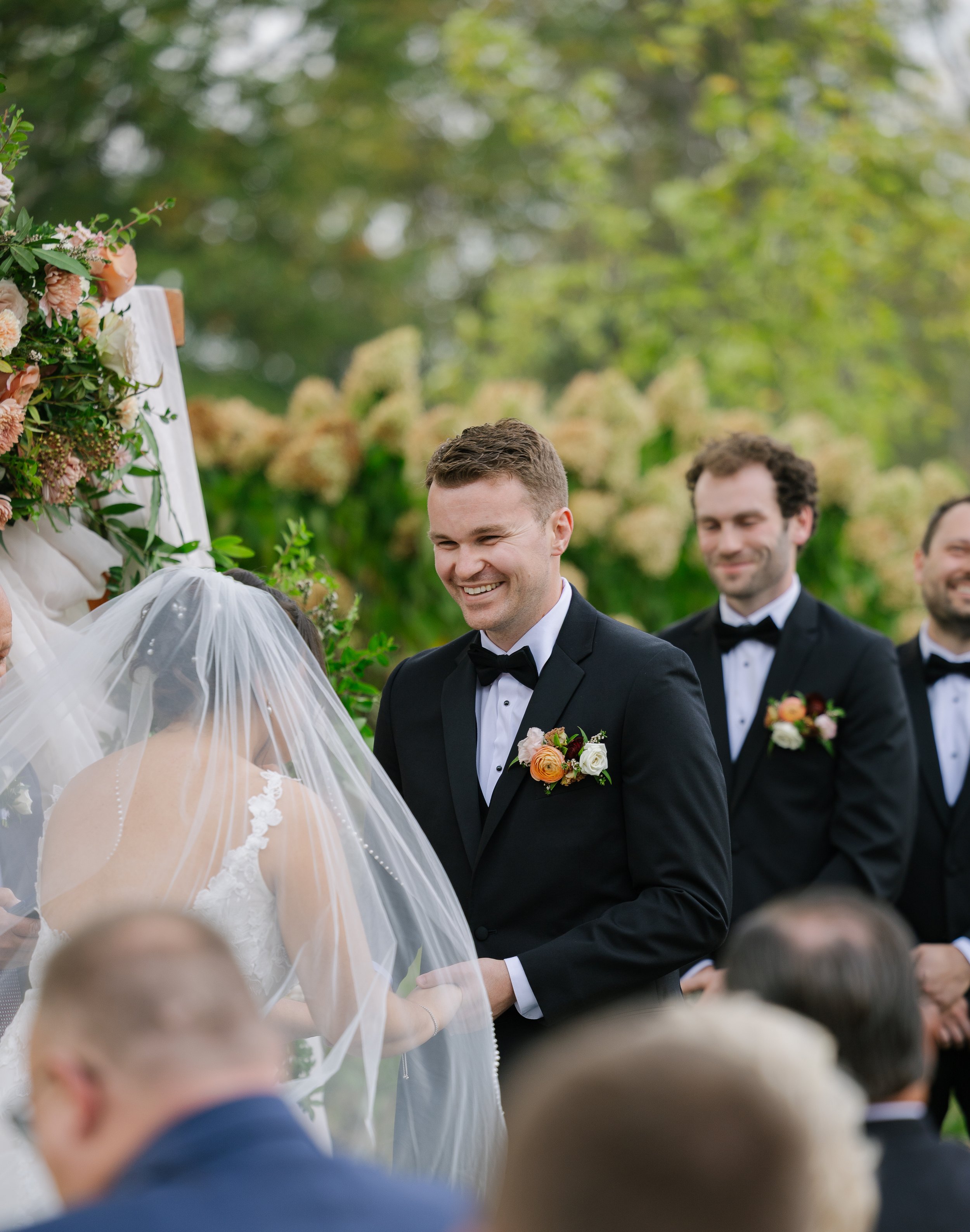 A groom smiling at a wedding ceremony outdoors, wearing a black tuxedo with a boutonniere, standing in front of the bride under a floral arch with blurred guests in the foreground.
