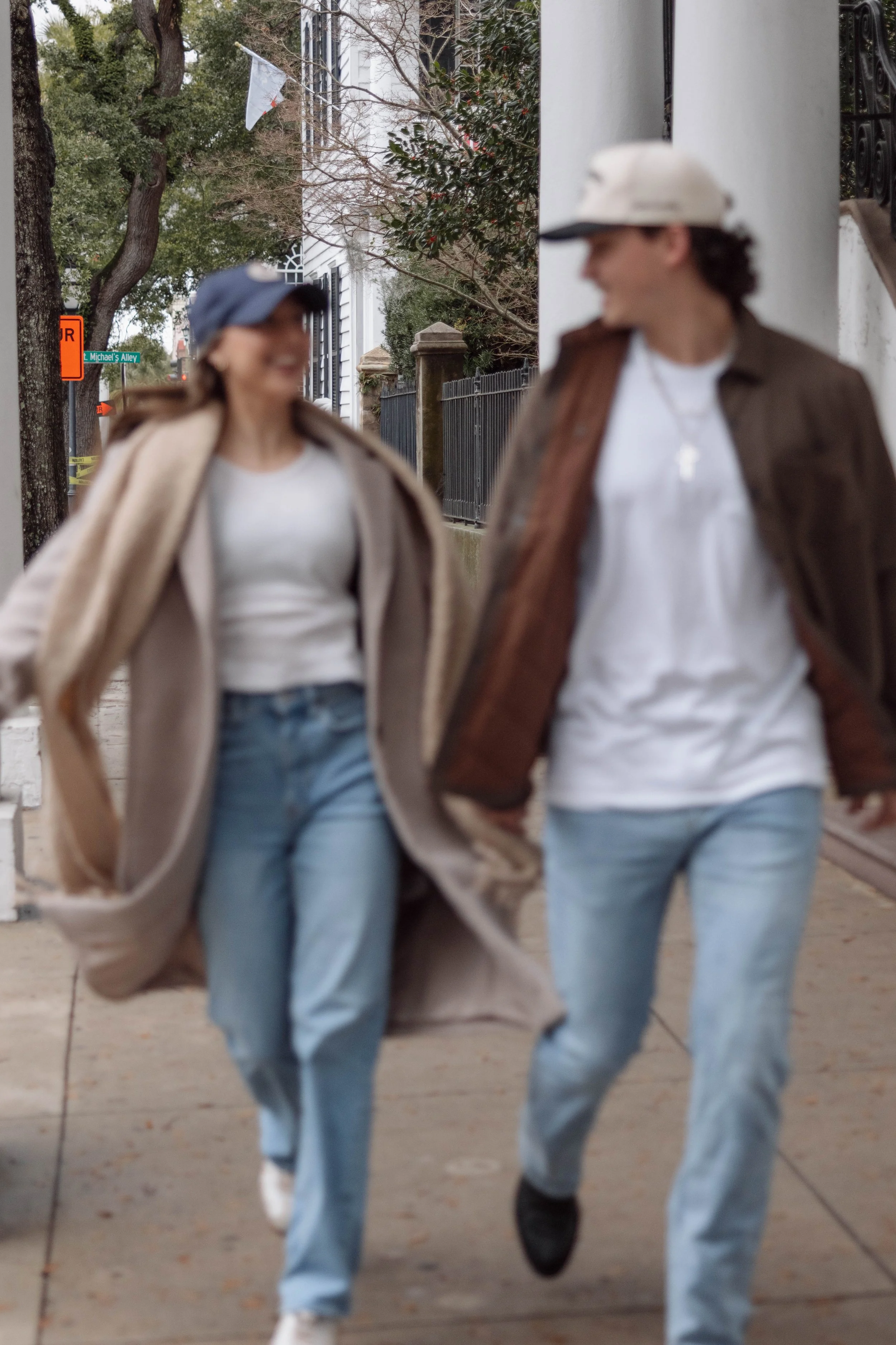 Two women walking on the sidewalk, wearing casual clothing and hats, smiling and looking at each other.