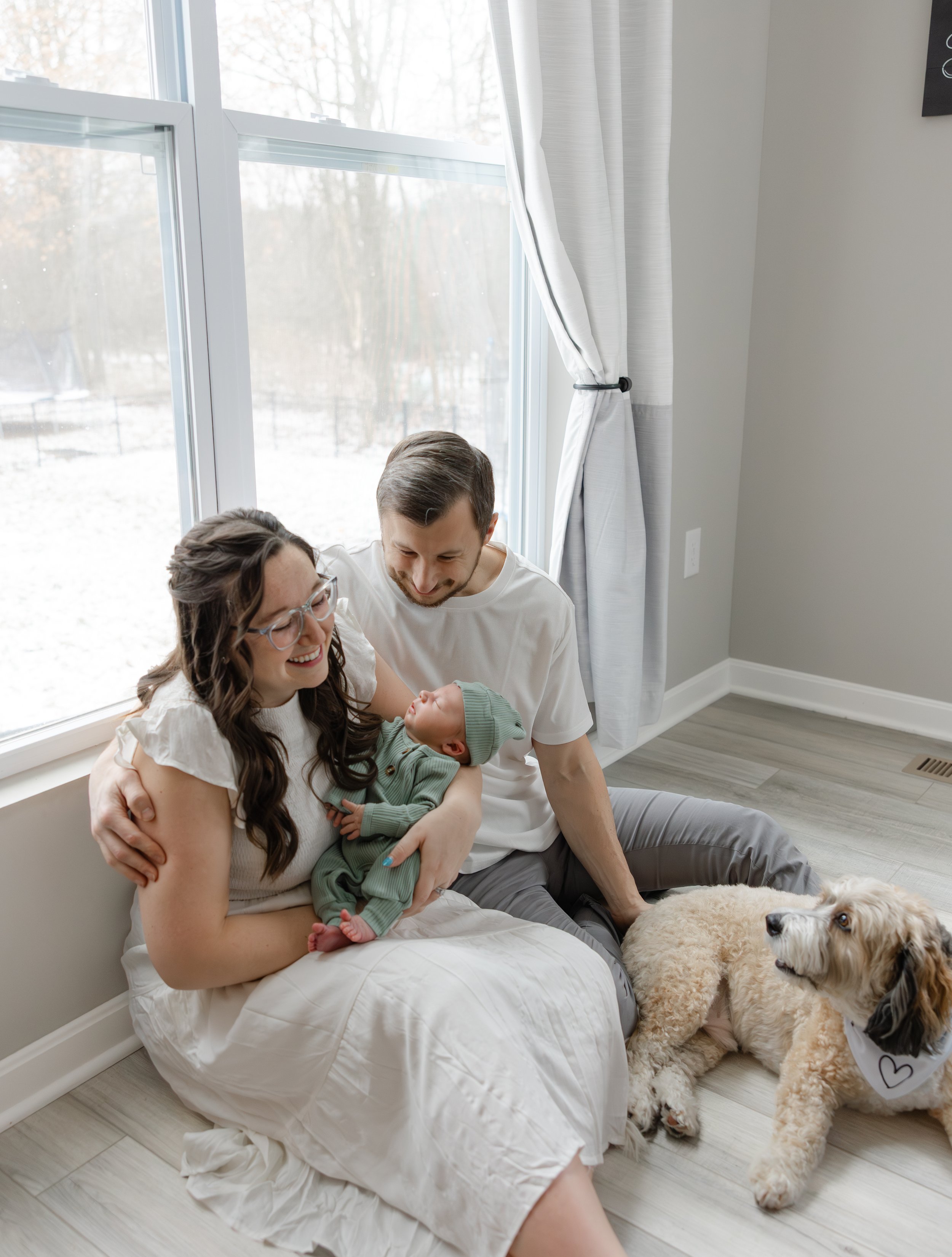 Family of three with a dog near a window, family members smiling at a newborn baby.