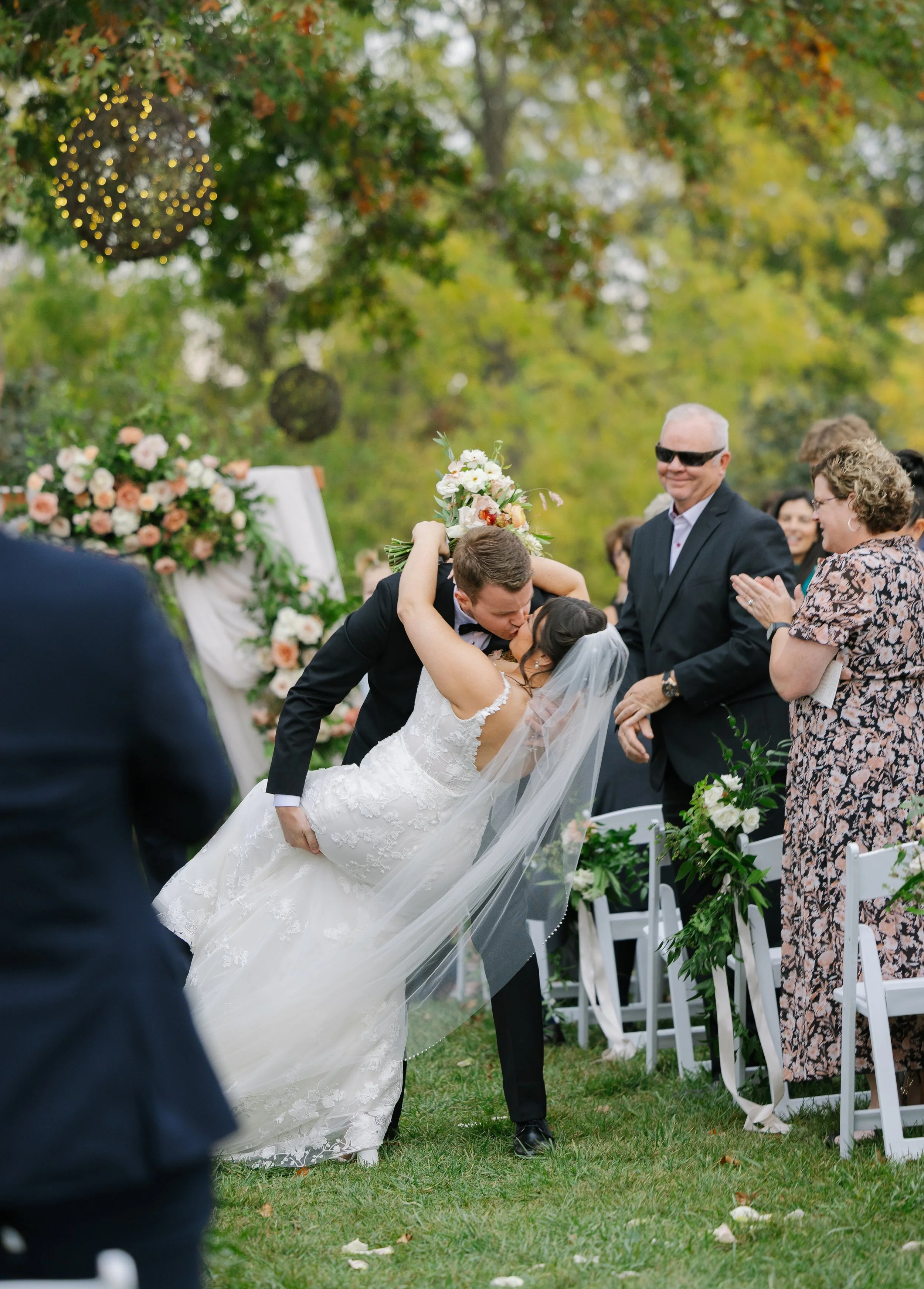 A newlywed couple kissing during their outdoor wedding ceremony, surrounded by friends and family, with floral decorations and an autumnal backdrop.