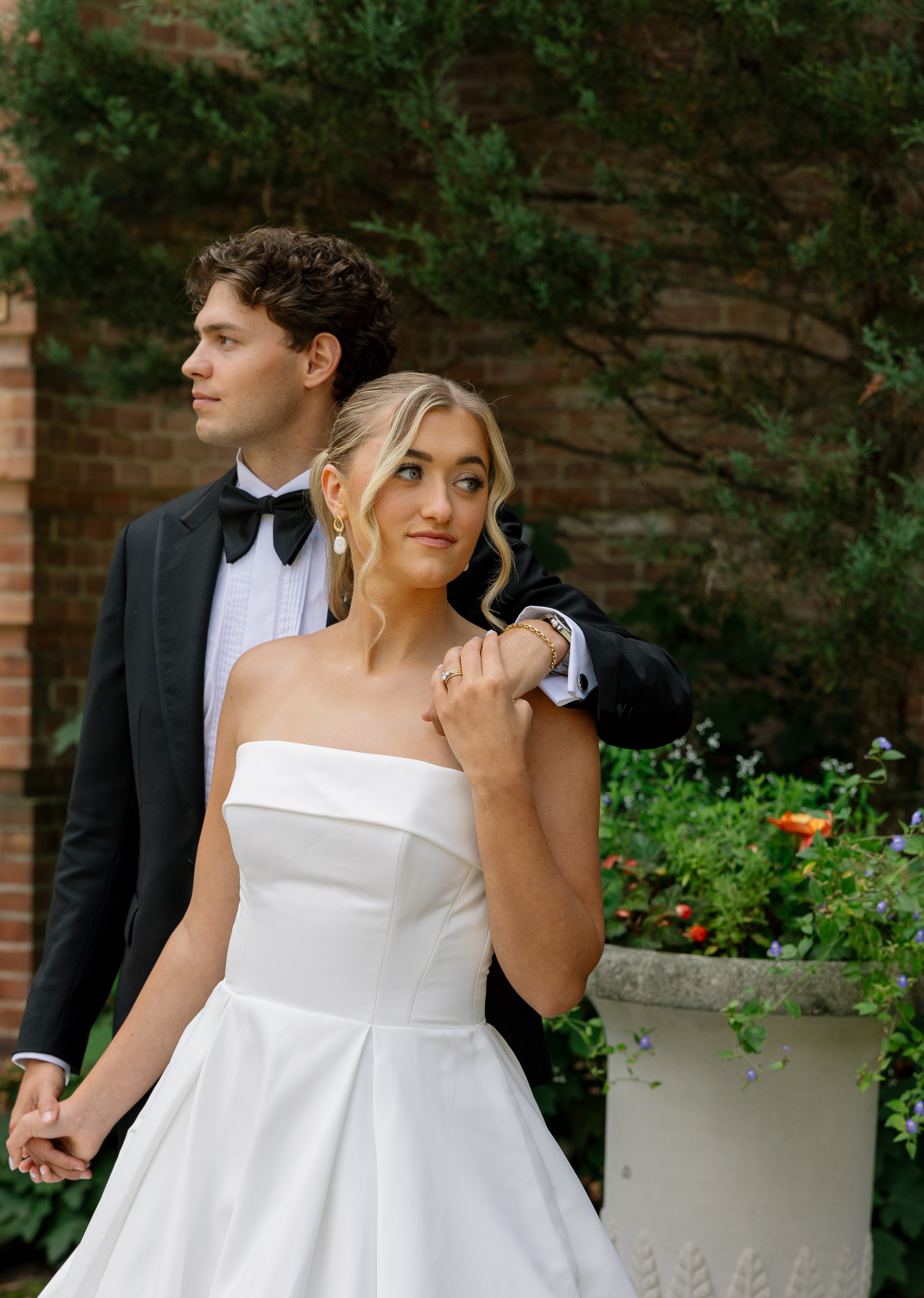 A bride and groom pose outside, with the groom standing behind the bride and resting his arm on her shoulder. The bride is wearing a strapless white wedding dress and the groom is in a black tuxedo with a bow tie. They are holding hands, and there are plants and flowers in the background.