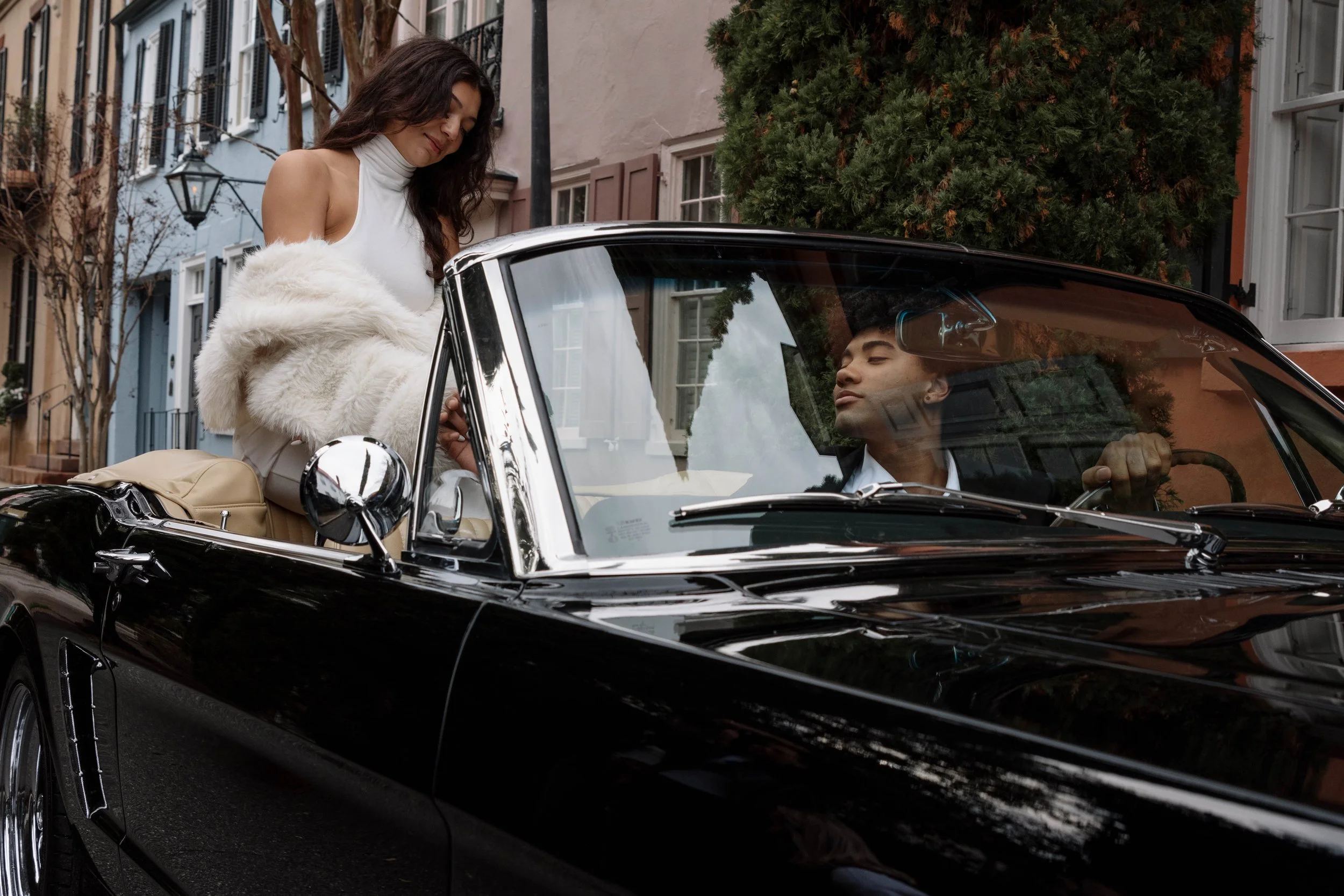 A woman in a white sleeveless top and beige pants is sitting on the back of a black vintage convertible car, holding a white faux fur jacket, as she interacts with a man in the driver's seat. The man appears to be looking at her, with a city street w