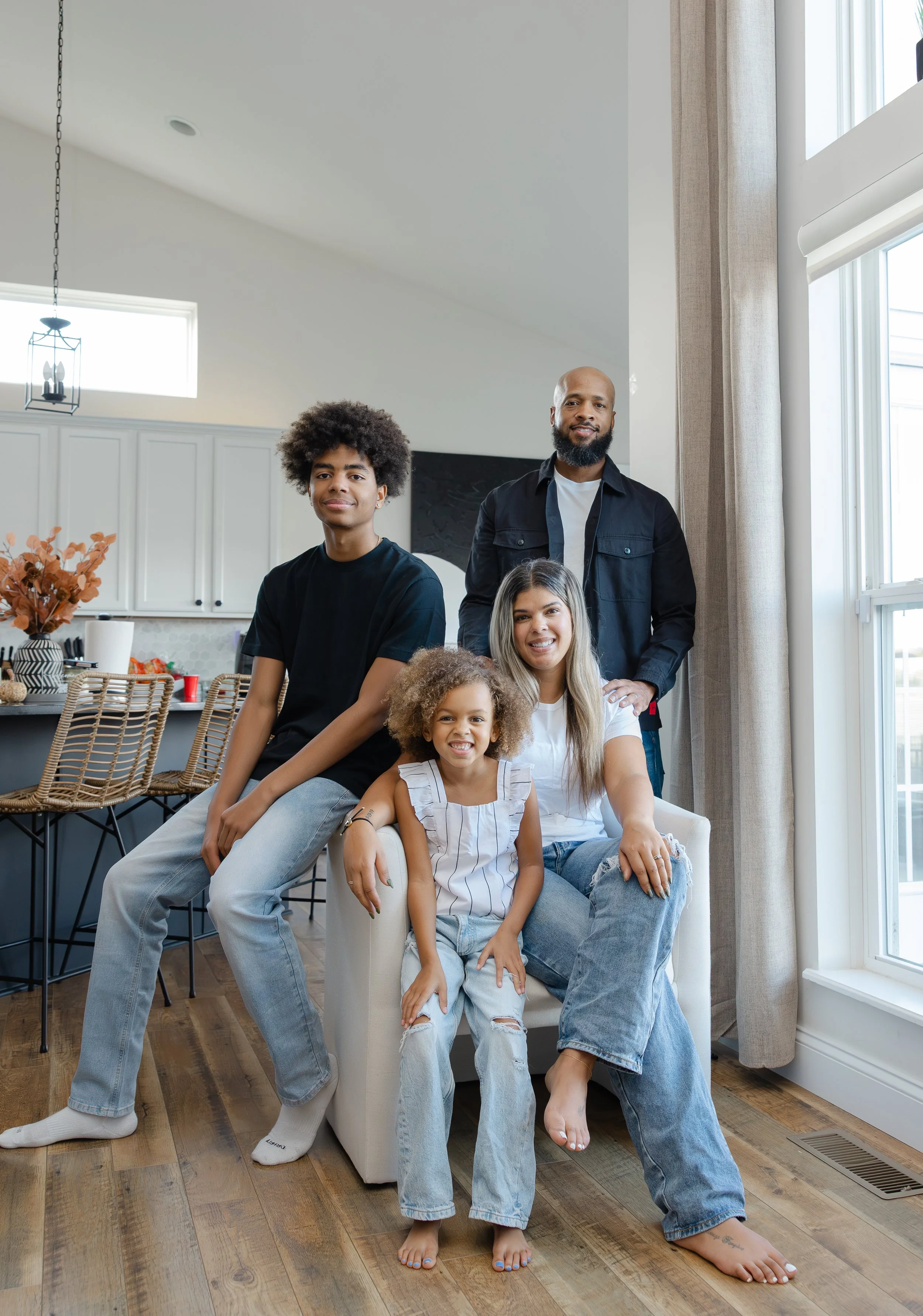 A family of five posing in a bright living room with hardwood floors, white walls, and large windows.