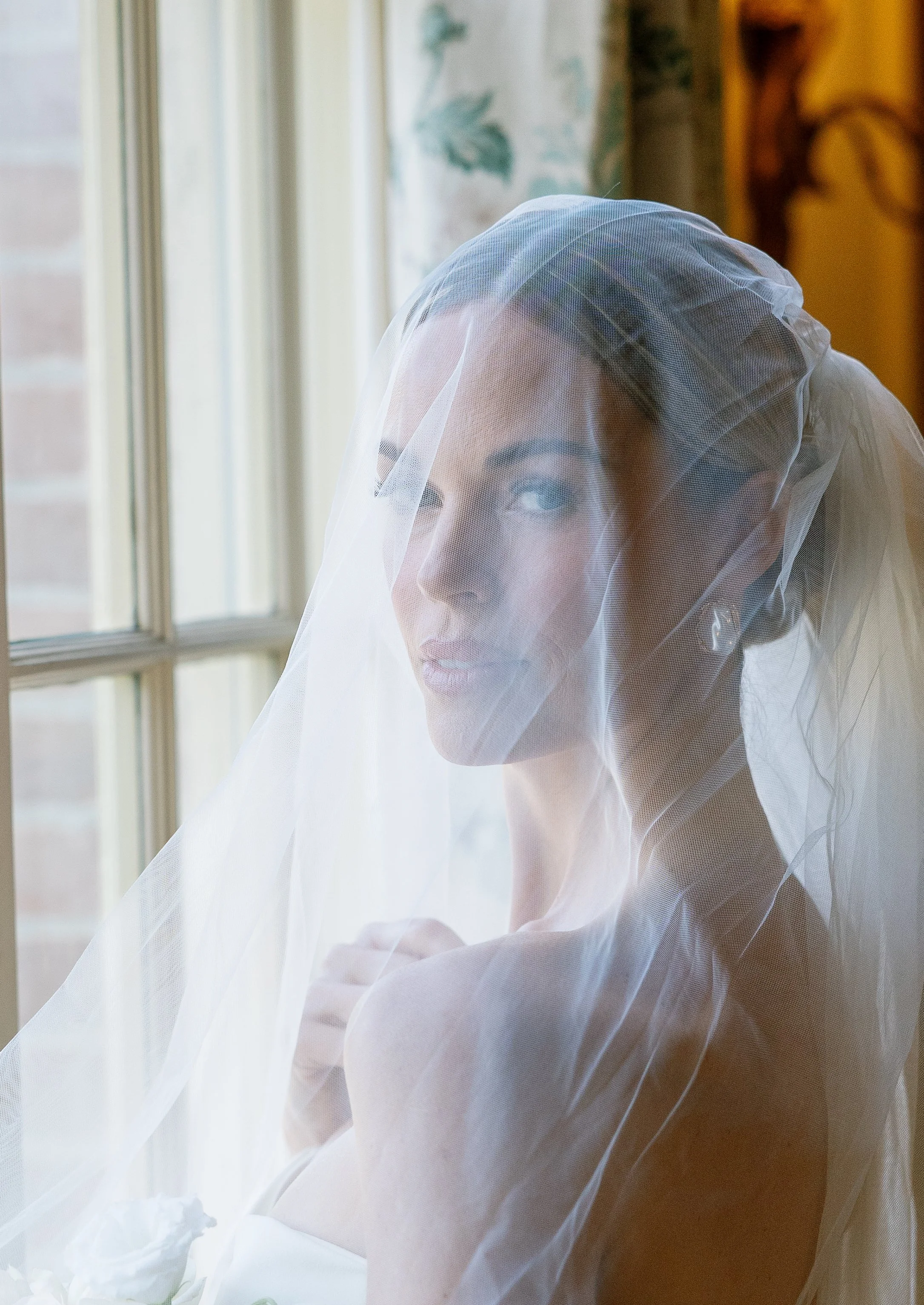 A woman with short hair and makeup wearing a wedding veil, looking slightly over her shoulder by a window.