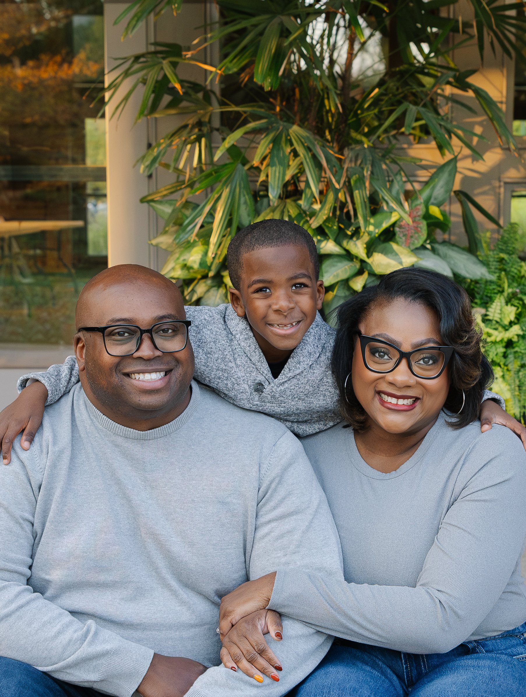 A smiling family of three posing outdoors, with a man on the left, woman on the right, and boy in the middle leaning on their shoulders, in front of a green leafy background.