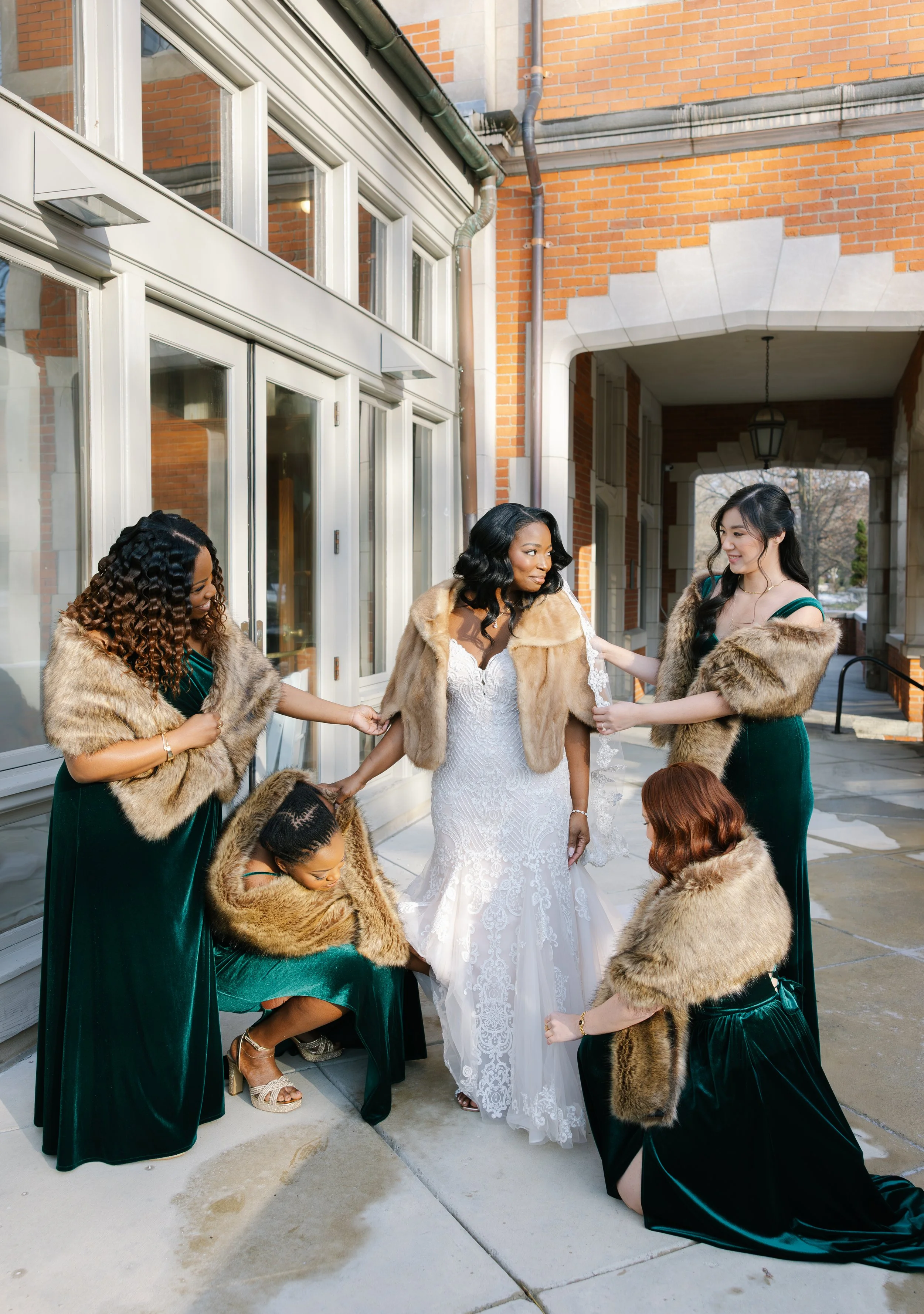 A bride in a white wedding gown and fur shawls surrounded by five bridesmaids in dark green dresses and fur stoles outside a brick building.