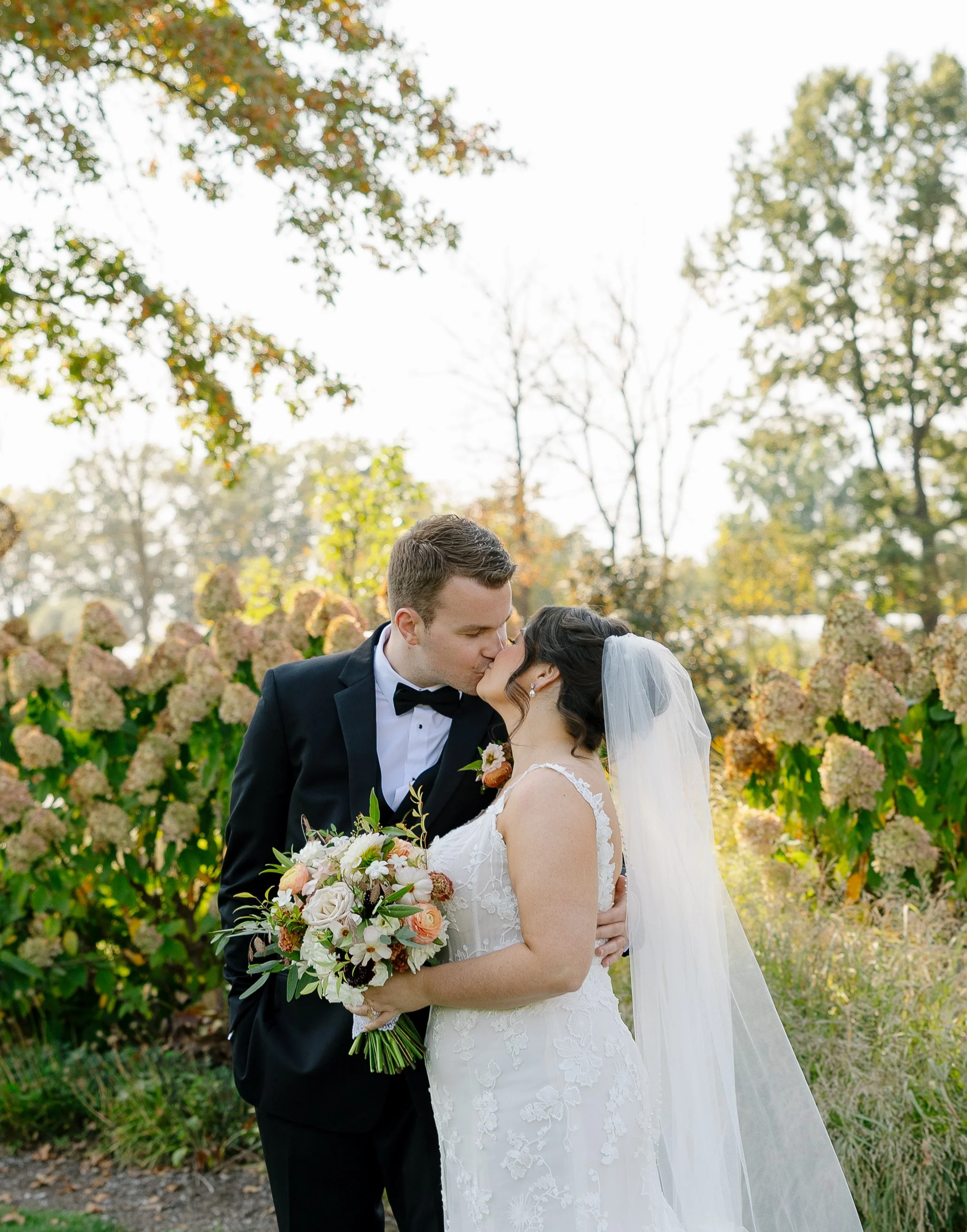 A newlywed couple sharing a kiss outdoors during daytime, surrounded by green and brown foliage, with the bride holding a bouquet of flowers.