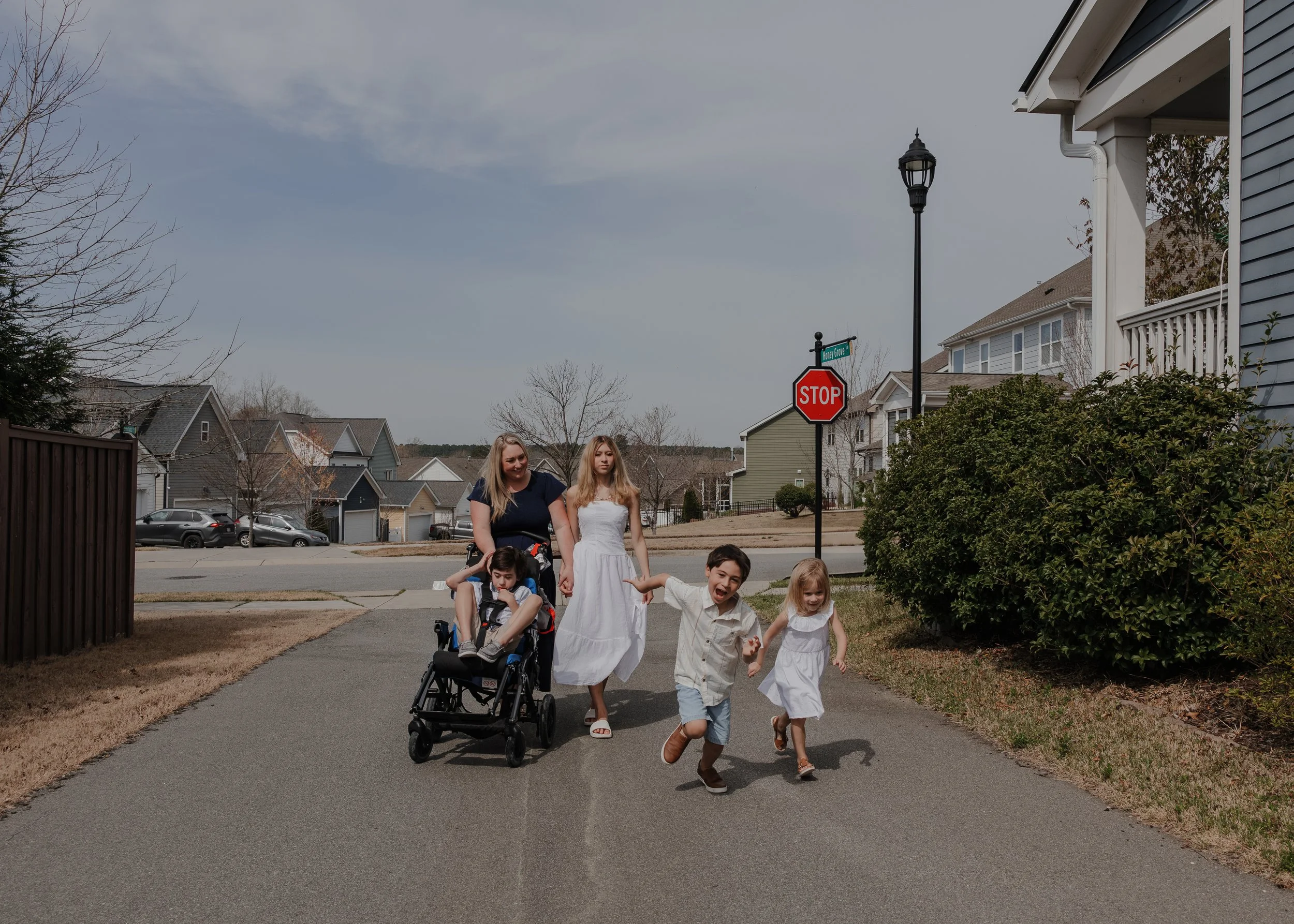 An inclusive family of six from Wendell, NC, walking down a residential street during a special needs photography session, with children on foot and in an adaptive mobility device.