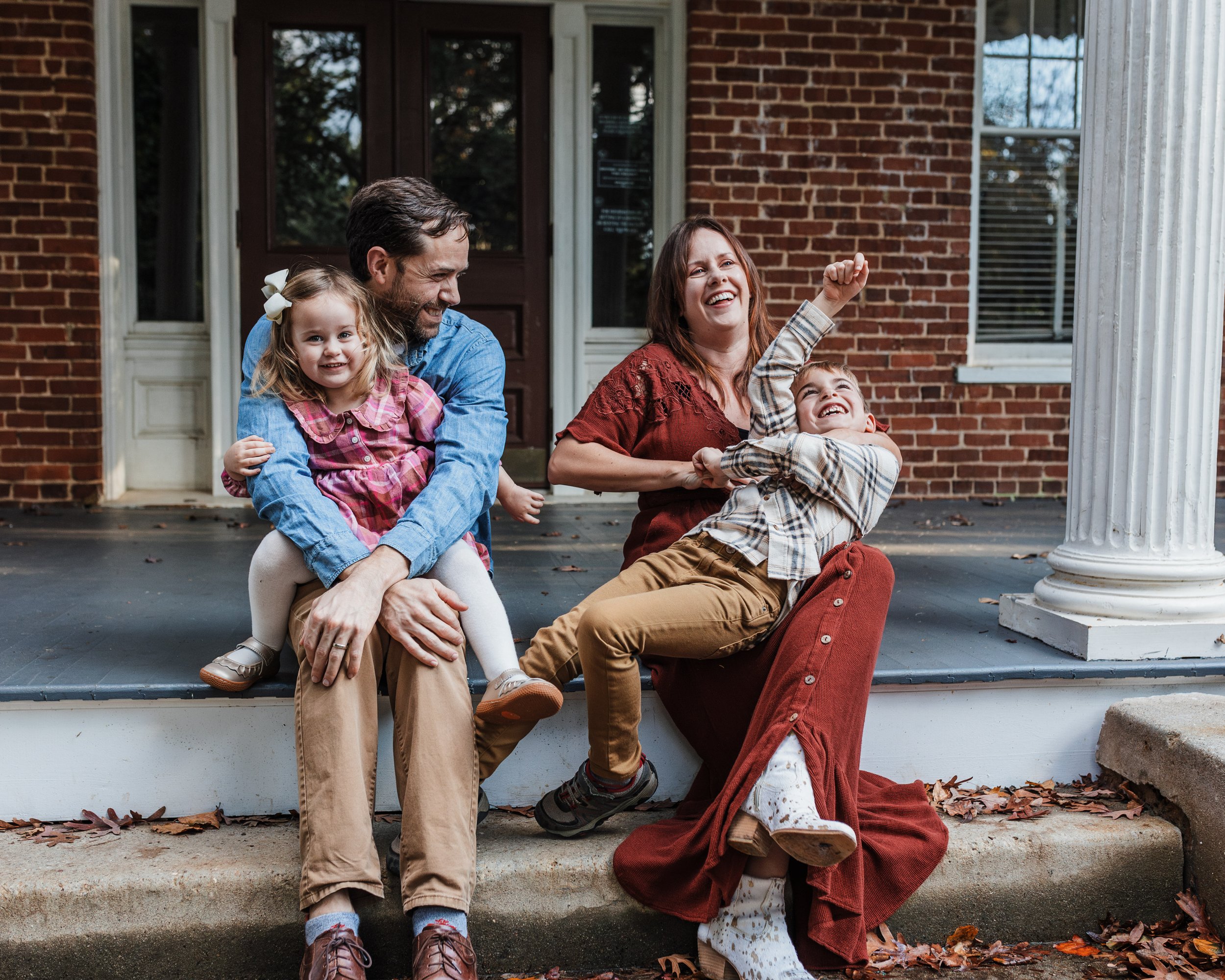 Candid family photo in Raleigh with parents sitting on a front porch at Fred Fletcher Park laughing with their two young children, capturing playful connection and real family interaction outdoors