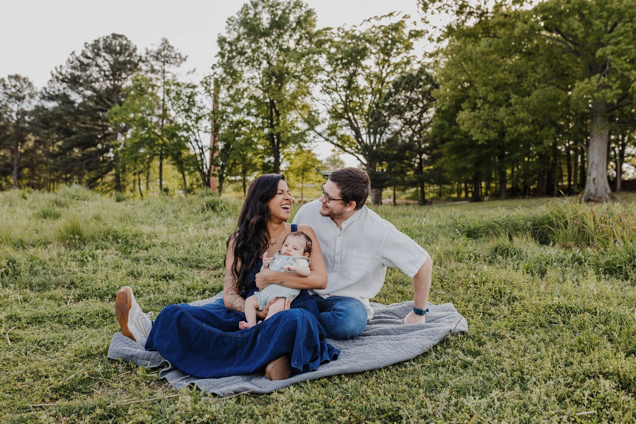 Wife and husband laugh with their baby during a Raleigh newborn photography session at the North Carolina Museum of Art.