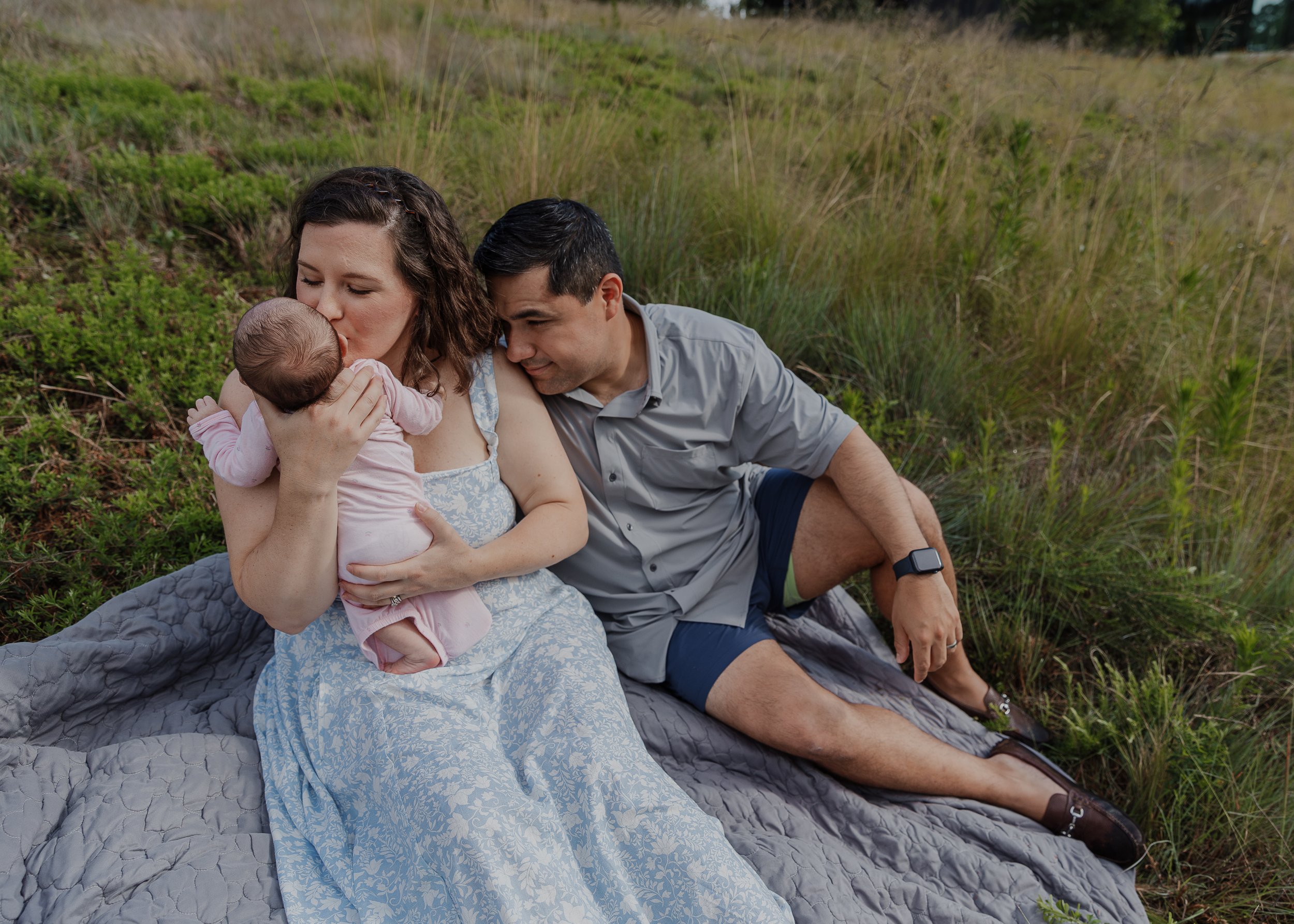 Mom and dad sit and snuggle baby during newborn photo session outside in Raleigh, NC.