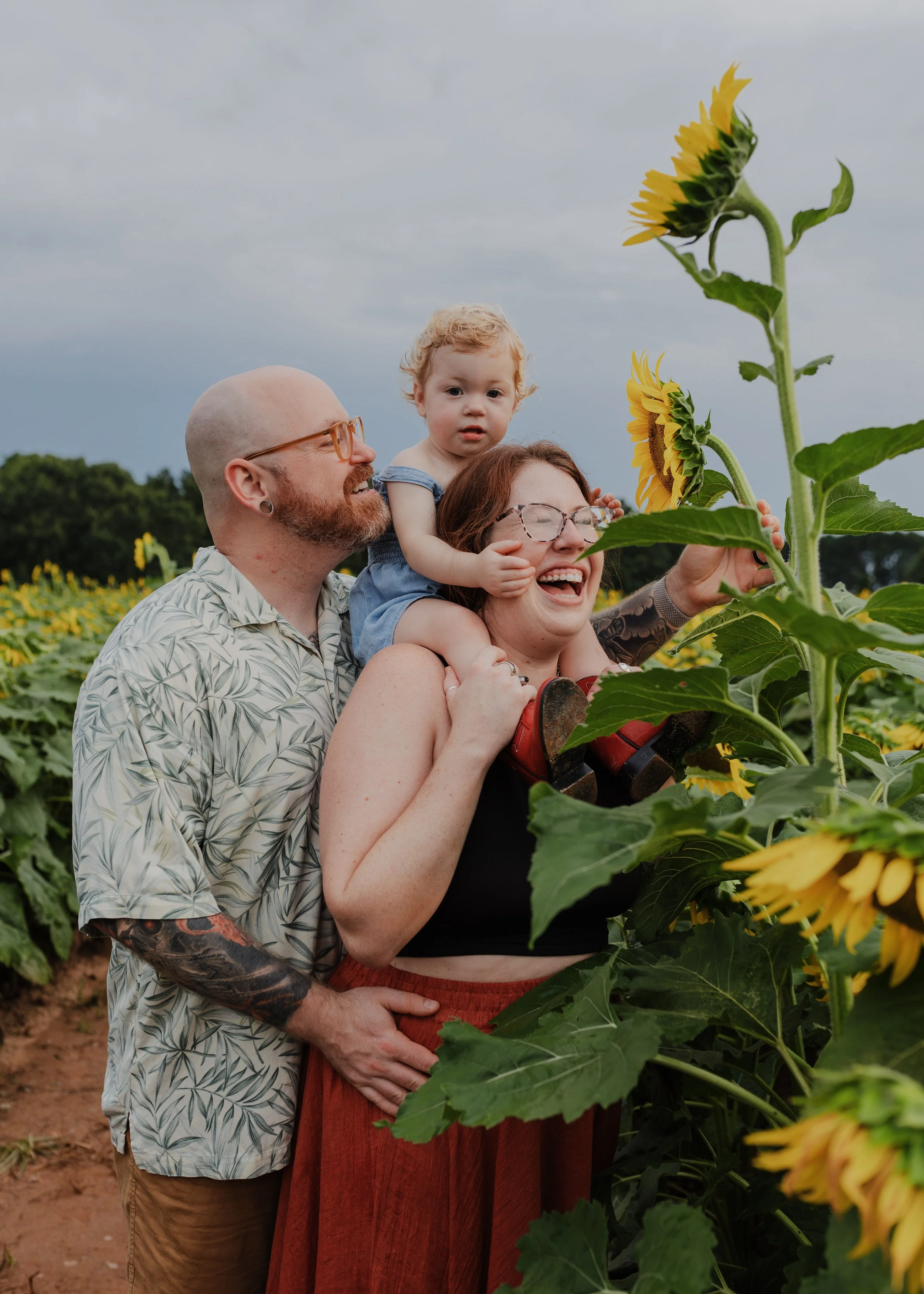Family with toddler laugh in a sunflower field at Dix Park in Raleigh, NC.