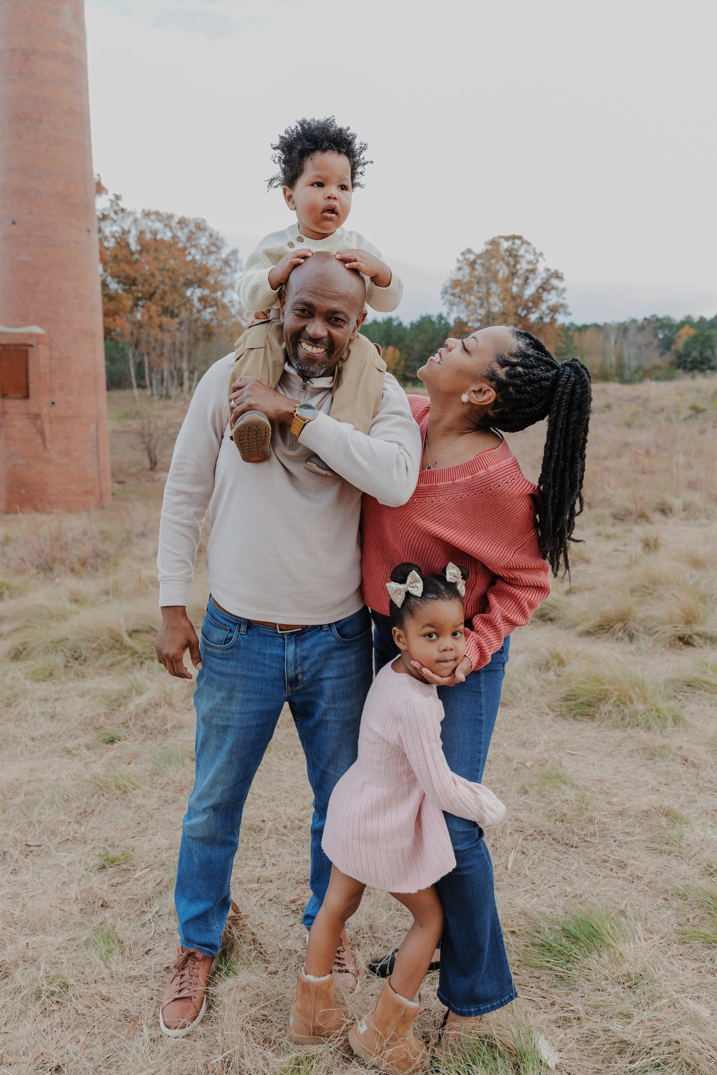 Baby rides on dad's shoulders while mom and daughter pose during a fall family photo session at the NCMA park in Raleigh, NC.