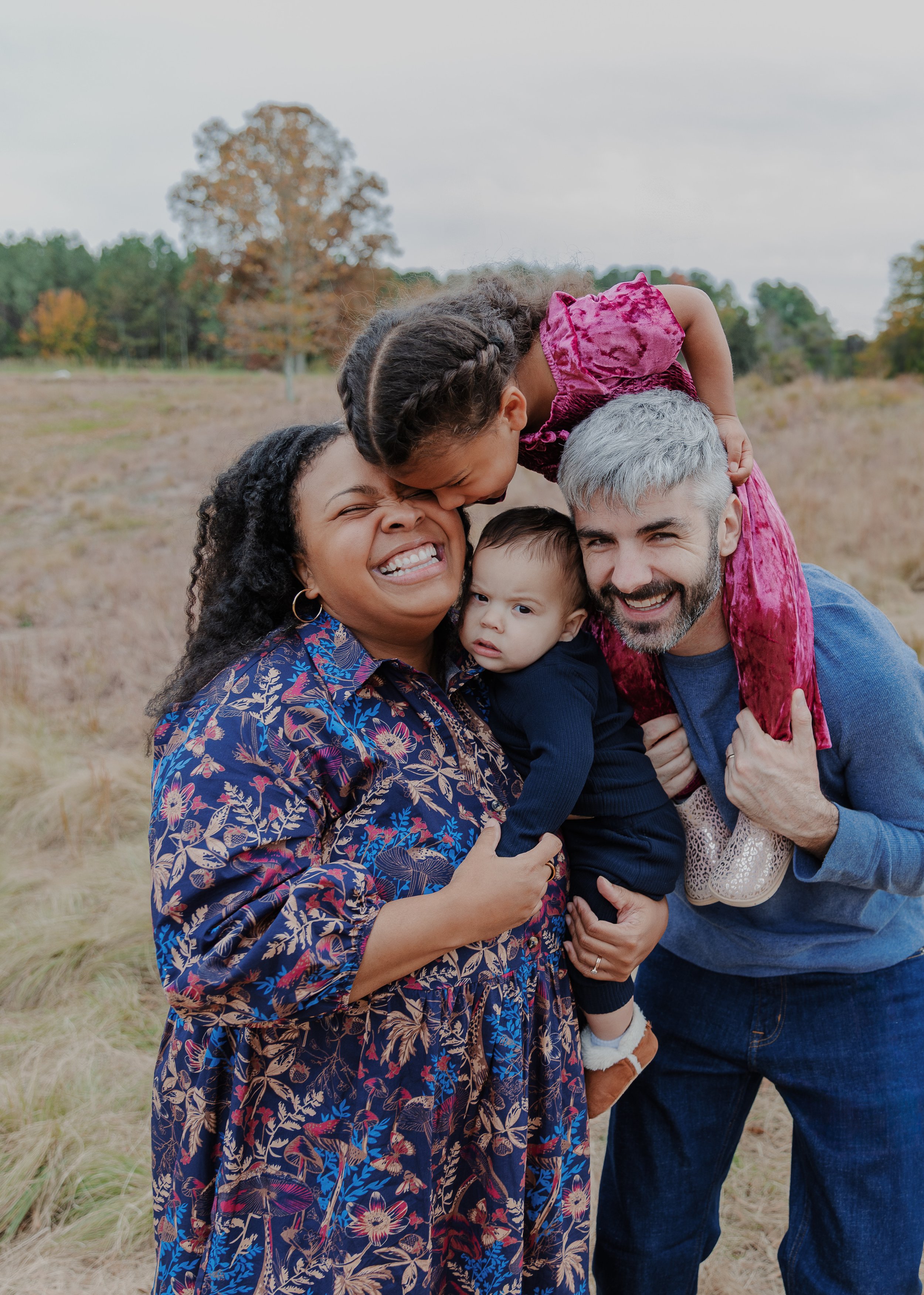 Mom and dad laugh with 3-year-old daughter and baby during a fall family mini session at the North Carolina Museum of Art Park in Raleigh.