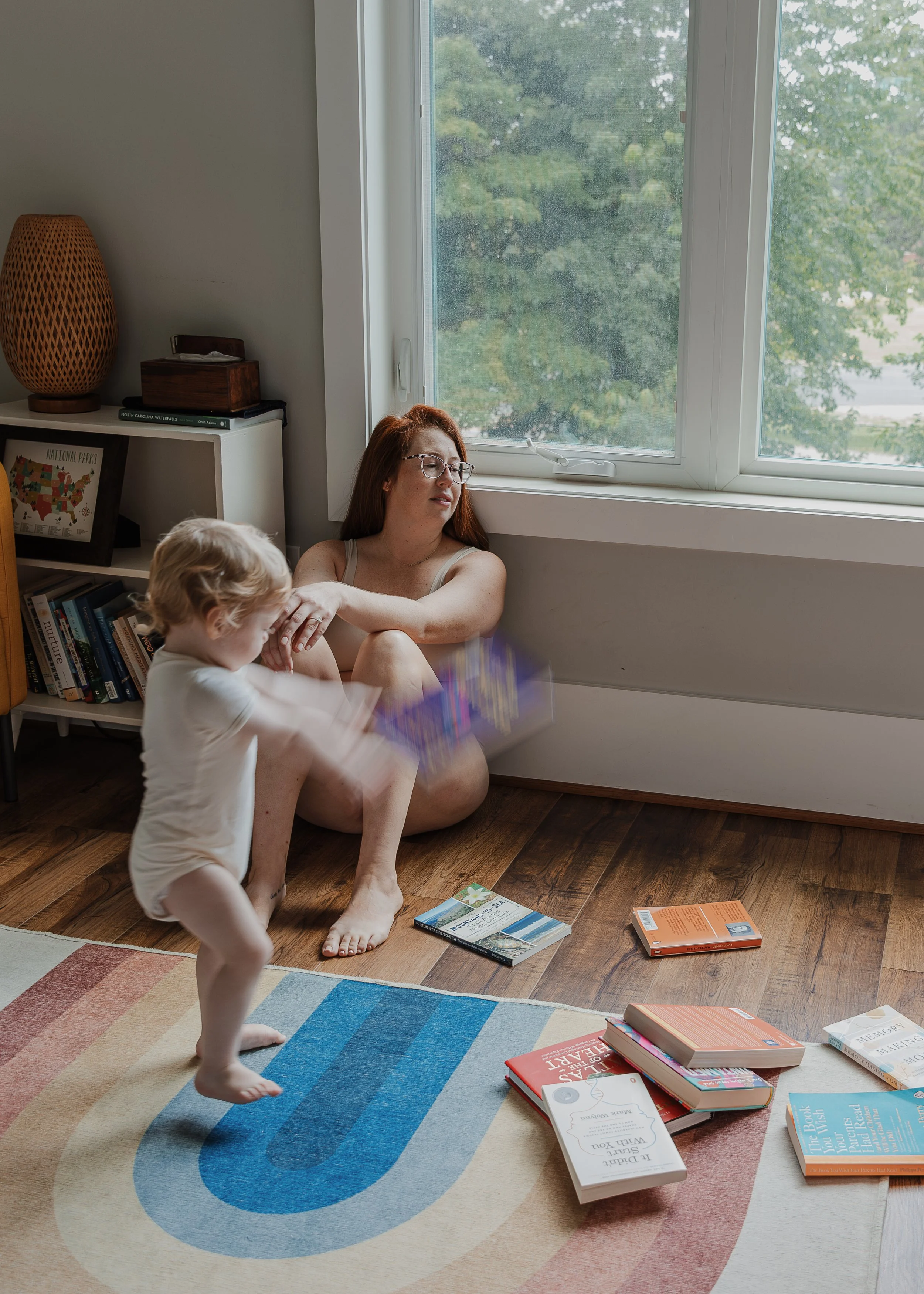 A mom leans back against a bedroom wall while her toddler throws books, captured during a motherhood session in Raleigh to show the chaos that comes with motherhood.
