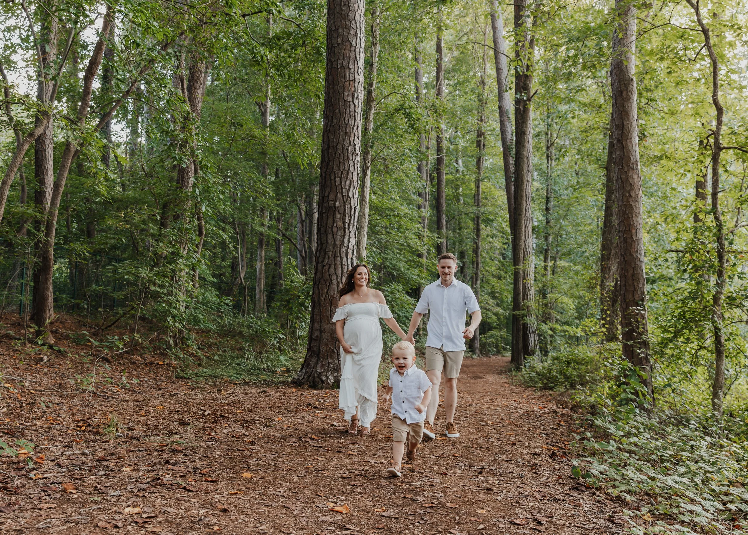 Pregnant woman holds her husband's hand and runs after her toddler during a maternity photo session at Historic Yates Mill County Park in Raleigh.