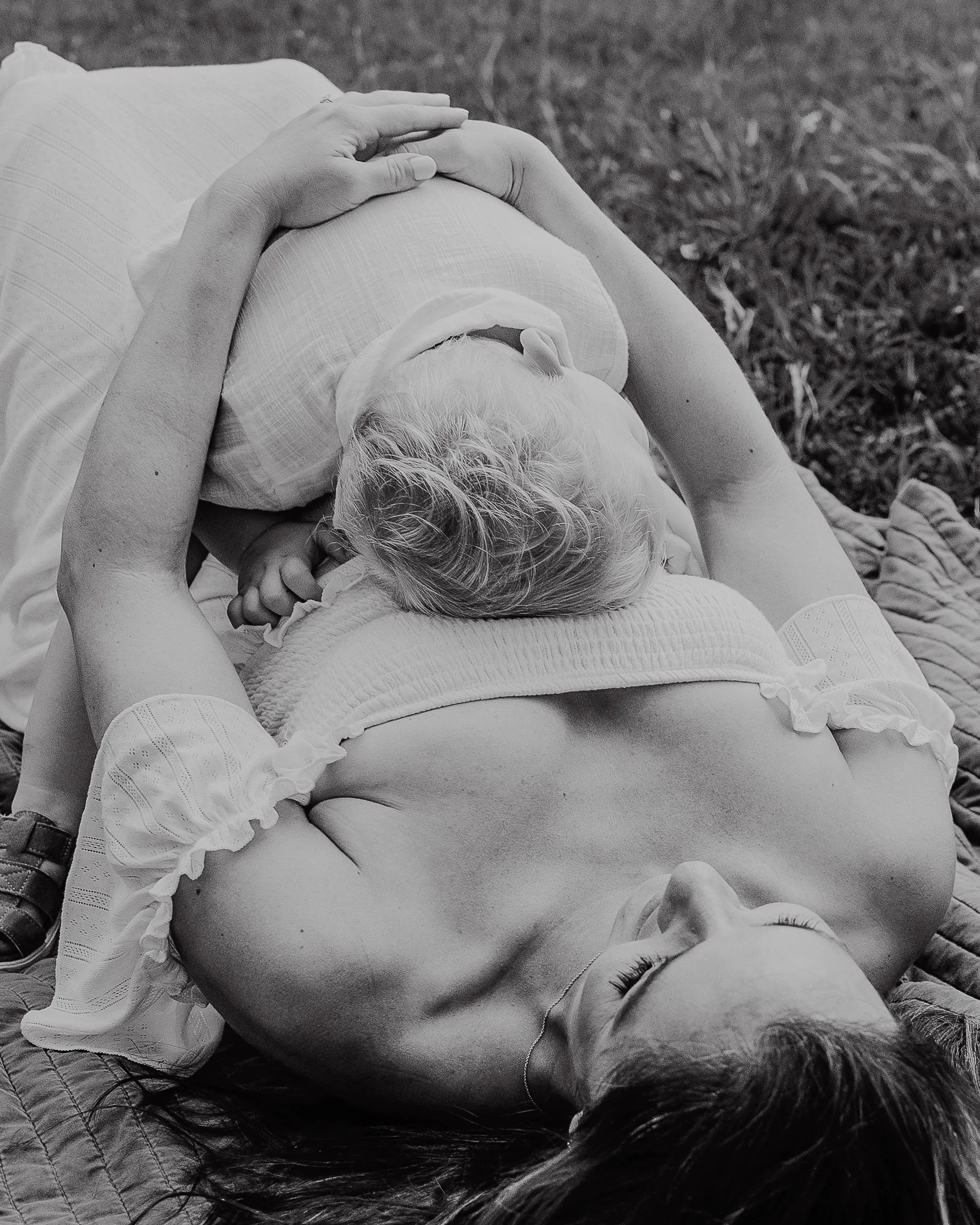 Black and white photo captured at a maternity photoshoot in Raleigh of a pregnant woman laying in the grass with her toddler hugging her belly.