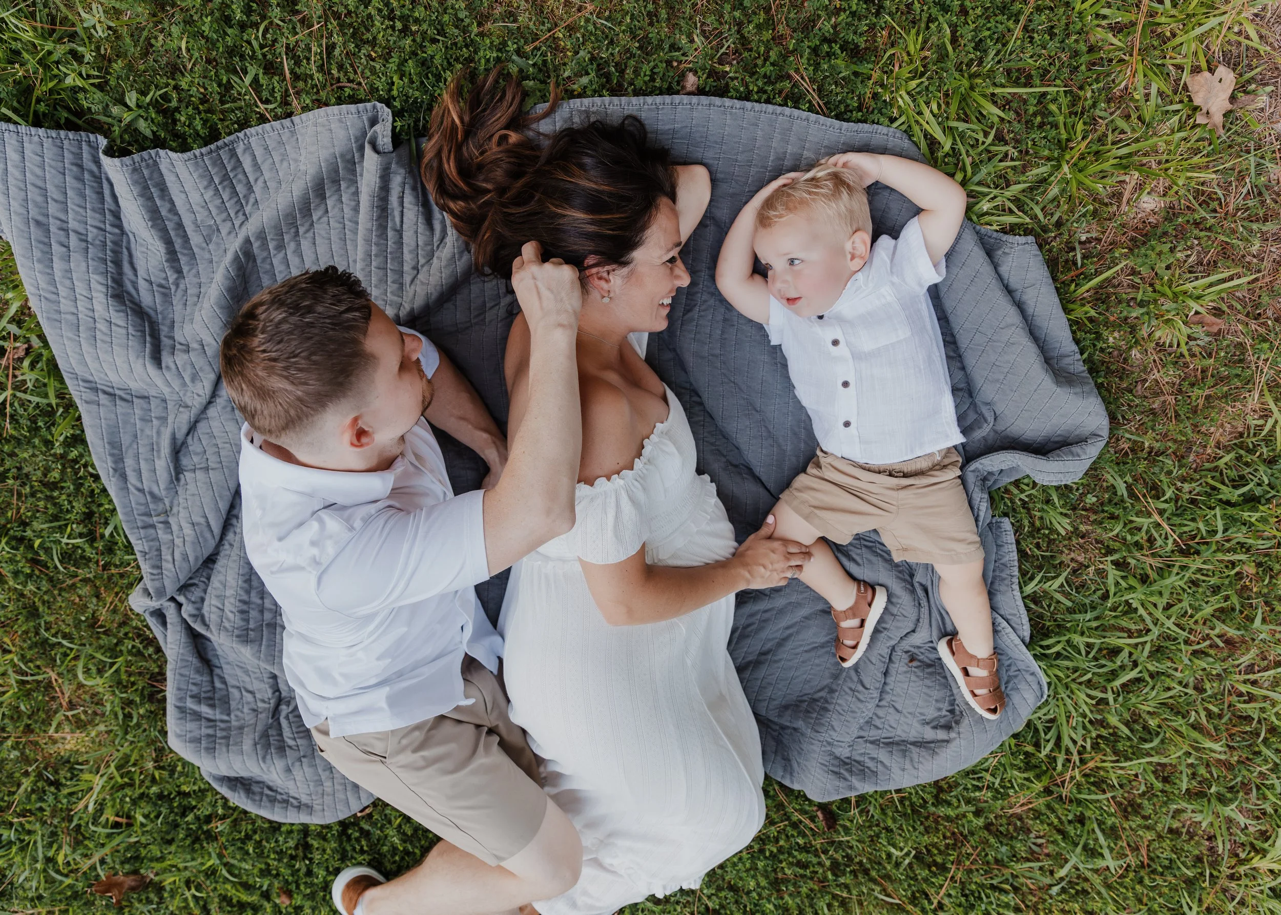 Pregnant woman and her husband lay with toddler son in the grass at Yates Mill County Park during a maternity photo session.