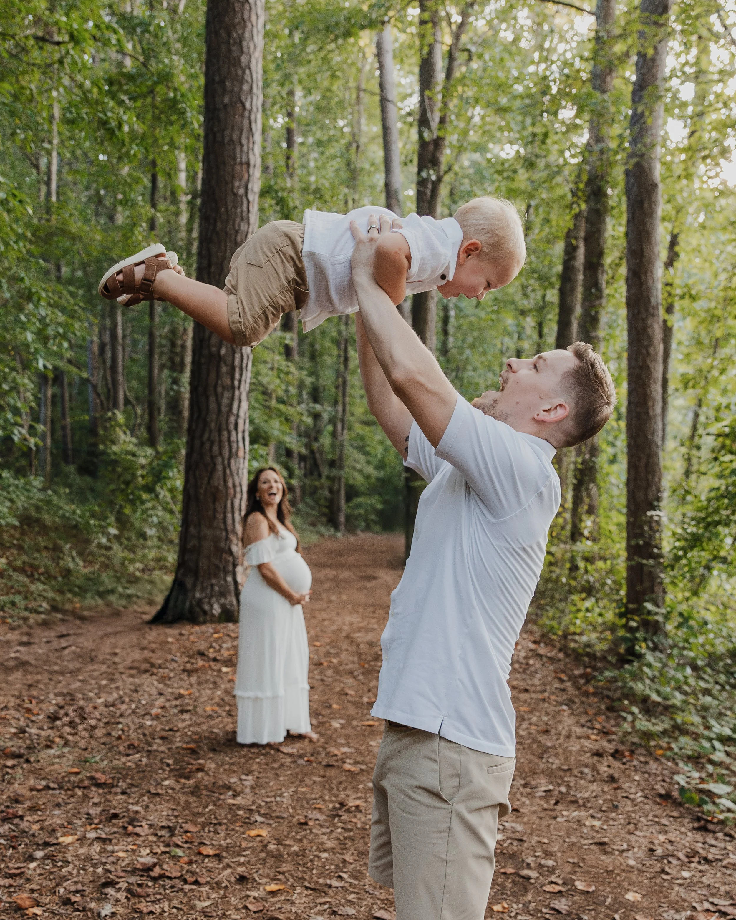 Dad throws toddler in the air while pregnant mom holds belly during a movement-focused maternity photo session at Historic Yates Mill County Park in Raleigh, NC.