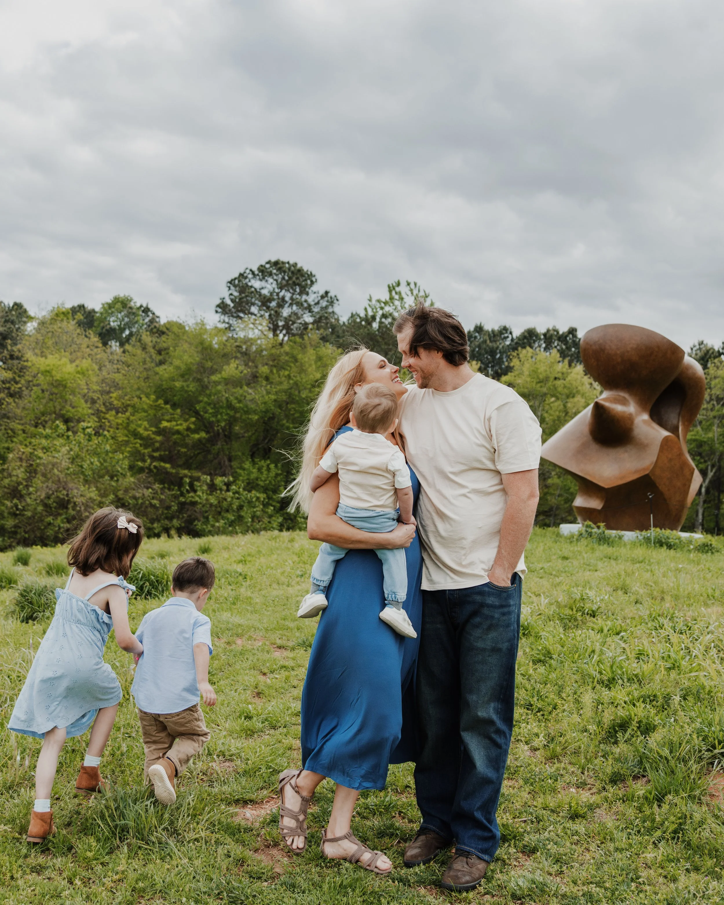Husband and wife hold baby and kiss while their 5-year-old daughter and 3-year-old son run in circles around them during spring mini sessions in Raleigh, NC.