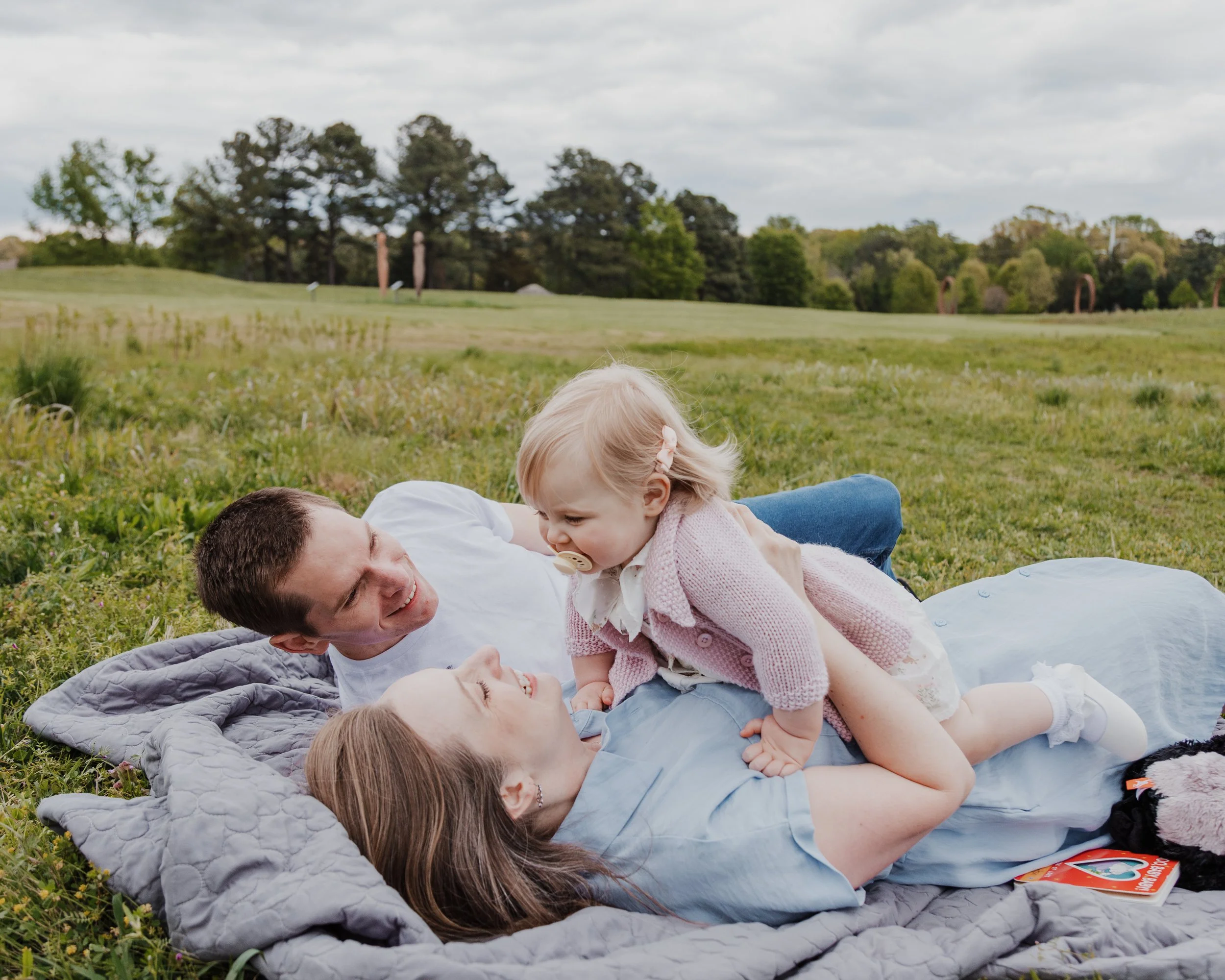 Husband and wife lay on ground and play with baby during a professional spring mini photo session at the NCMA park in Raleigh.