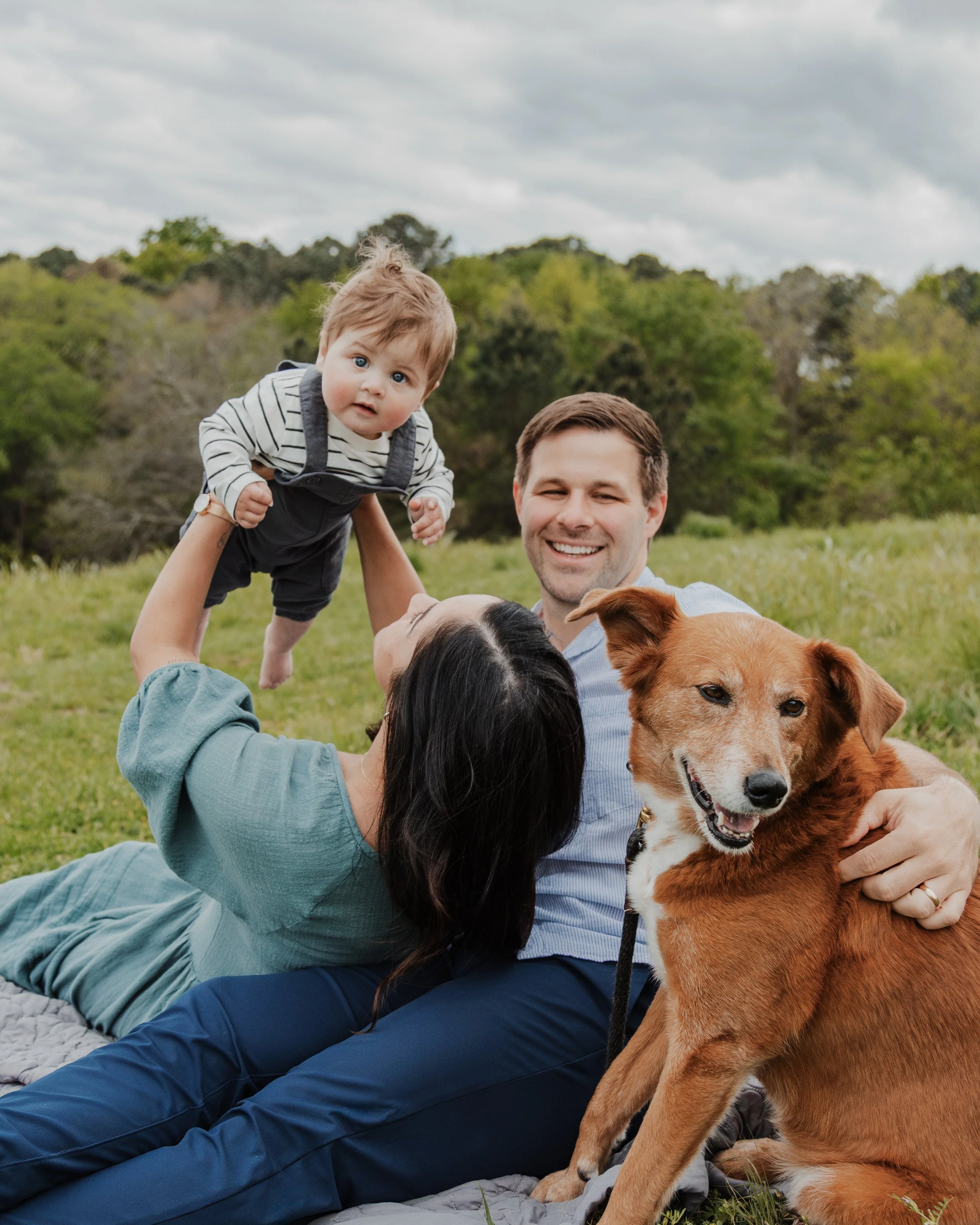 Husband and wife play with baby and dog during a spring mini session at the North Carolina Museum of Art in Raleigh, NC.