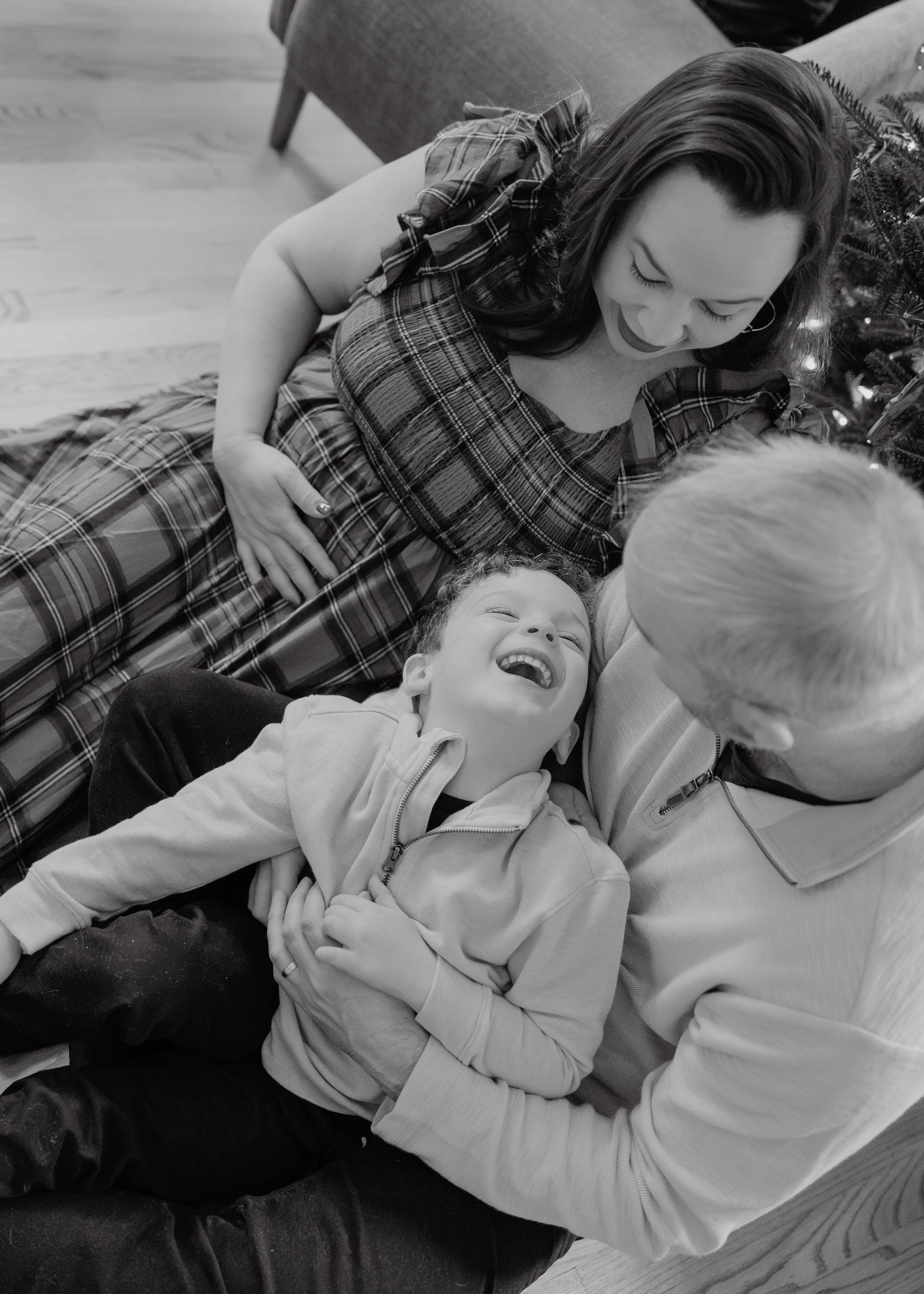 Mom, dad, and 4-year-old boy laugh in front of Christmas tree during a pregnancy photo session at their home in Durham, NC.