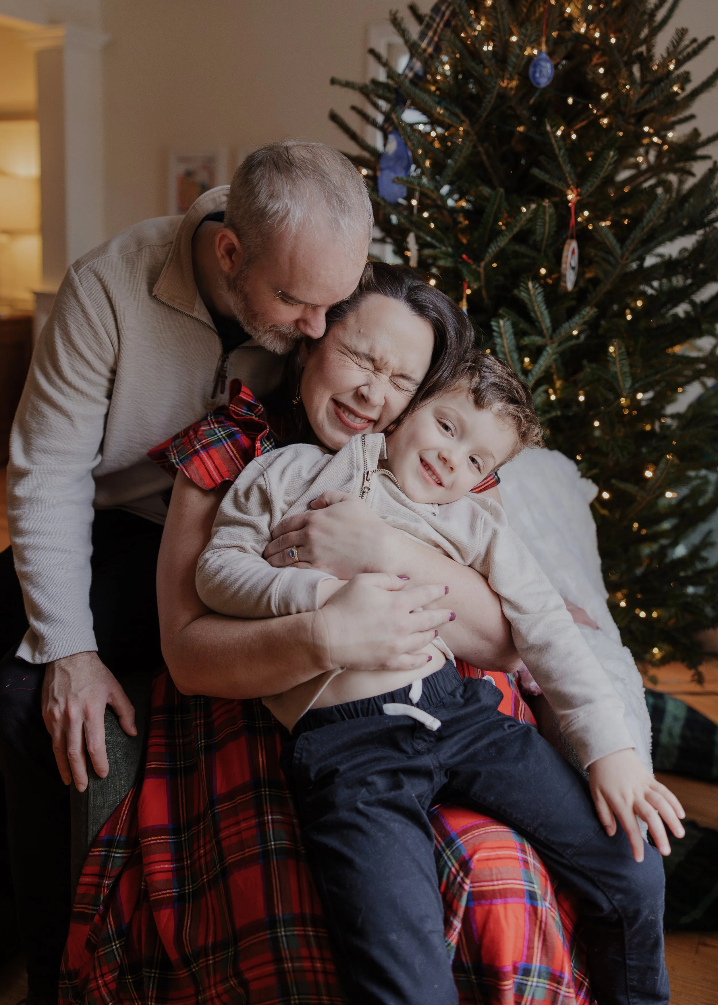 Mom, dad, and 4-year-old boy hugs on a chair in front of the Christmas tree at their home in Durham.
