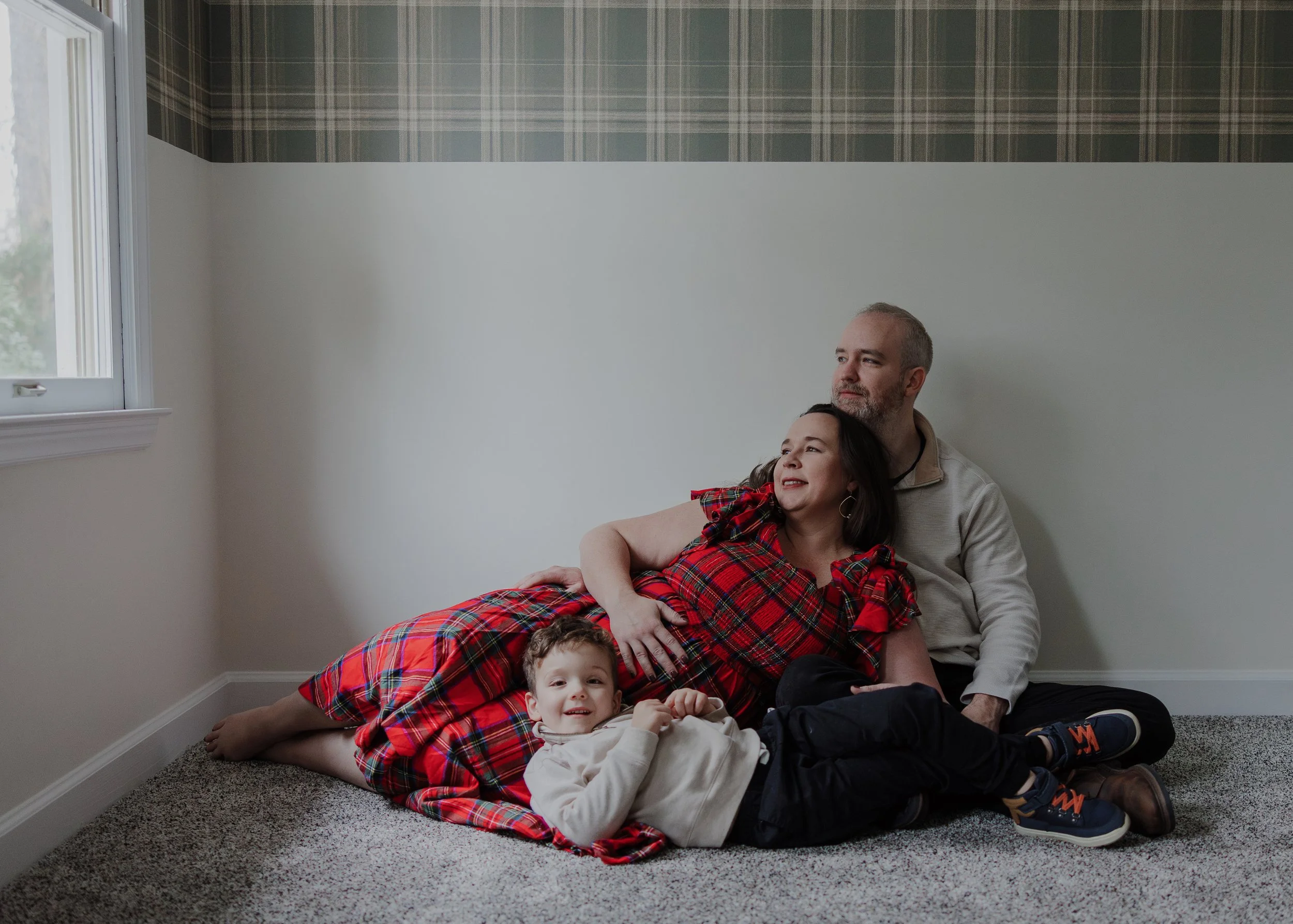 Pregnant mother in red plaid dress relaxing at home with partner and young child during an in-home maternity session by a Raleigh maternity photographer