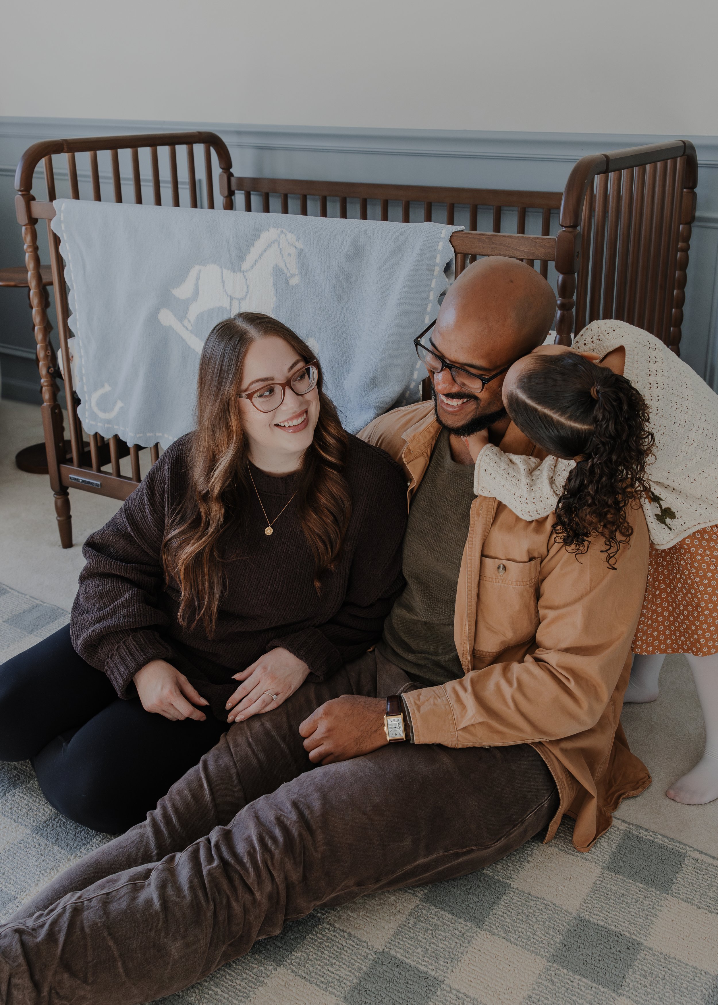 Mom, dad, and three year old girl play on the floor of new baby's nursery during a professional maternity photoshoot at their home in Wake Forest, NC.