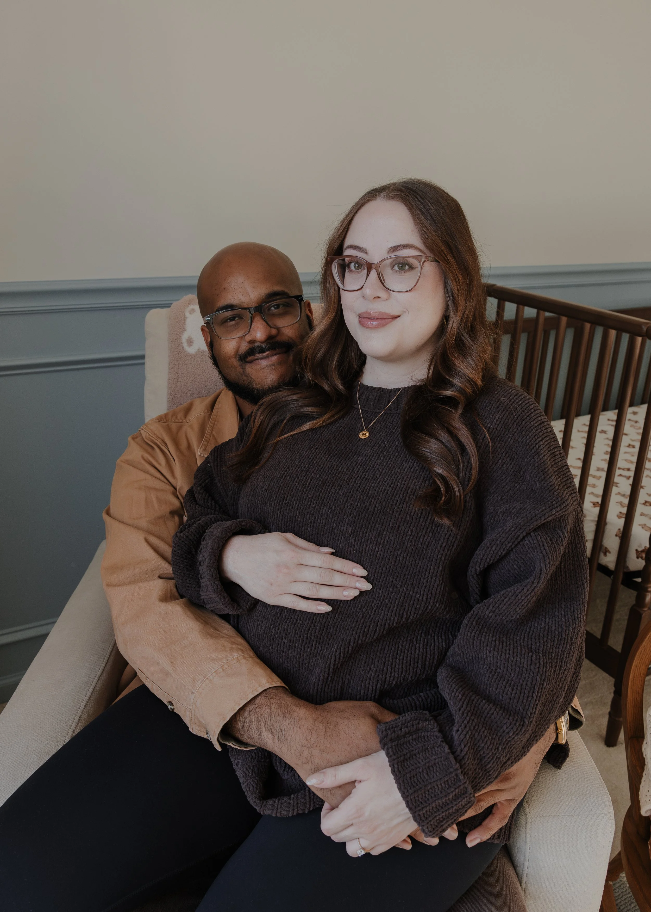 Pregnant woman sits on husband's lap in baby's nursery and smiles during a professional pregnancy photo session at their home in Wake Forest.