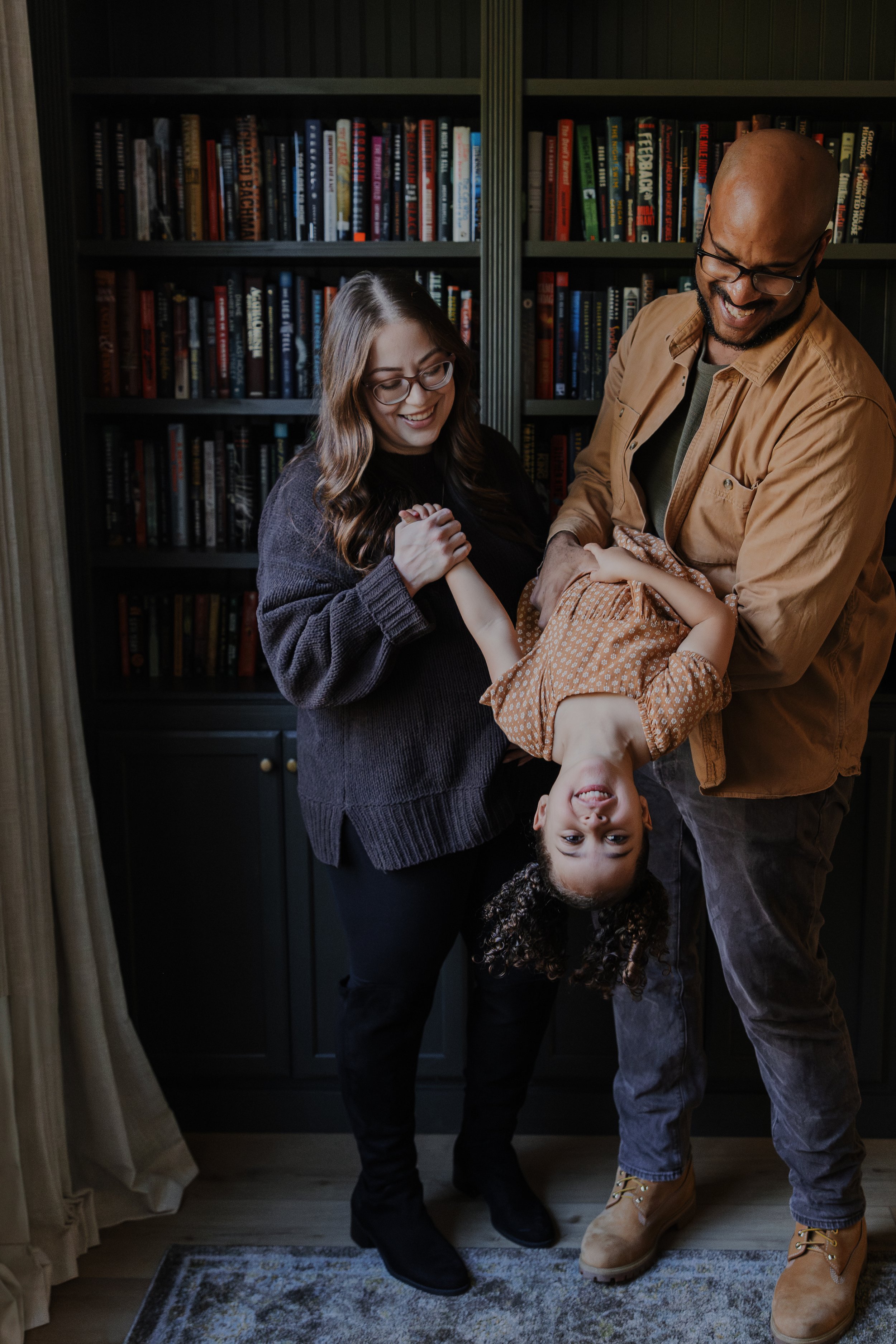 Dad holds three-year-old upside down in front of a built-in bookshelf while pregnant mom watches.