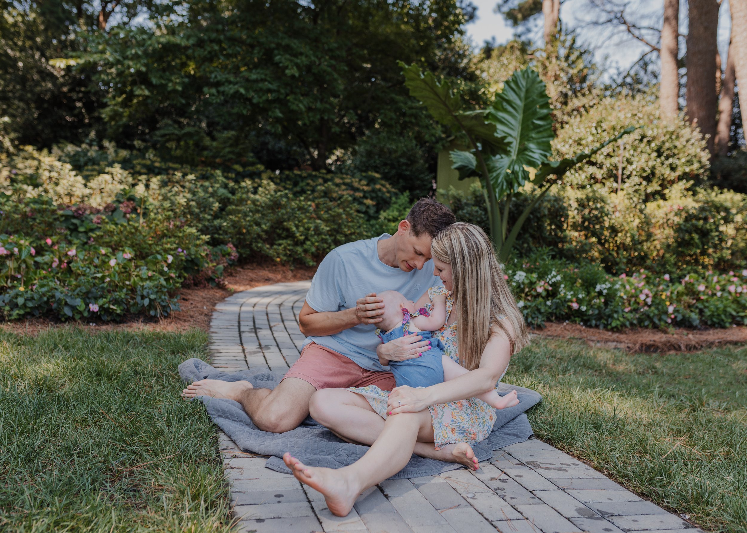Family with 6-month-old baby girl sit on a blanket in the WRAL Azalea Gardens during a spring family photo session