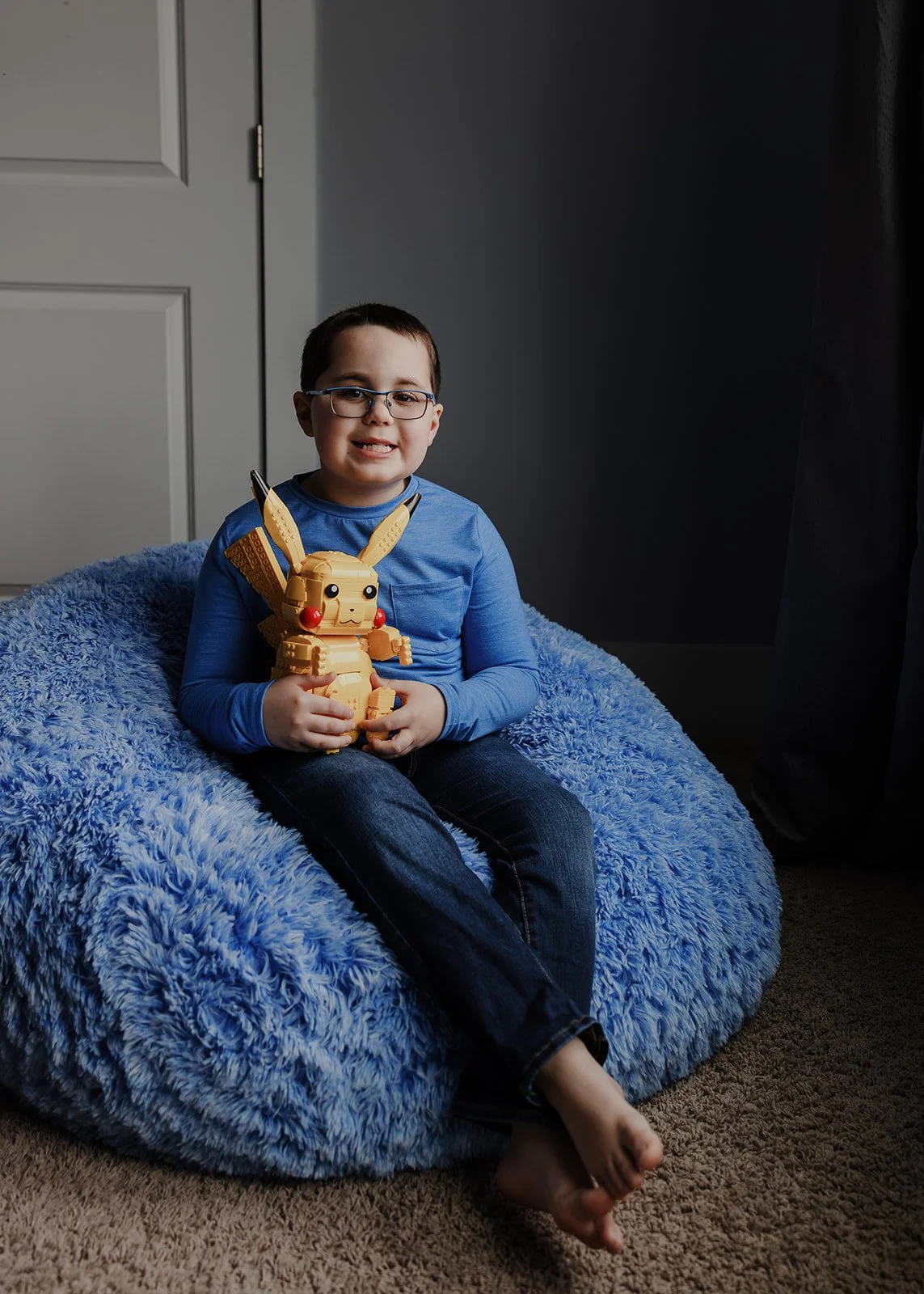 Child sitting on a blue bean bag holding a Pikachu LEGO build in his bedroom during an in-home family session in Wake Forest, NC.