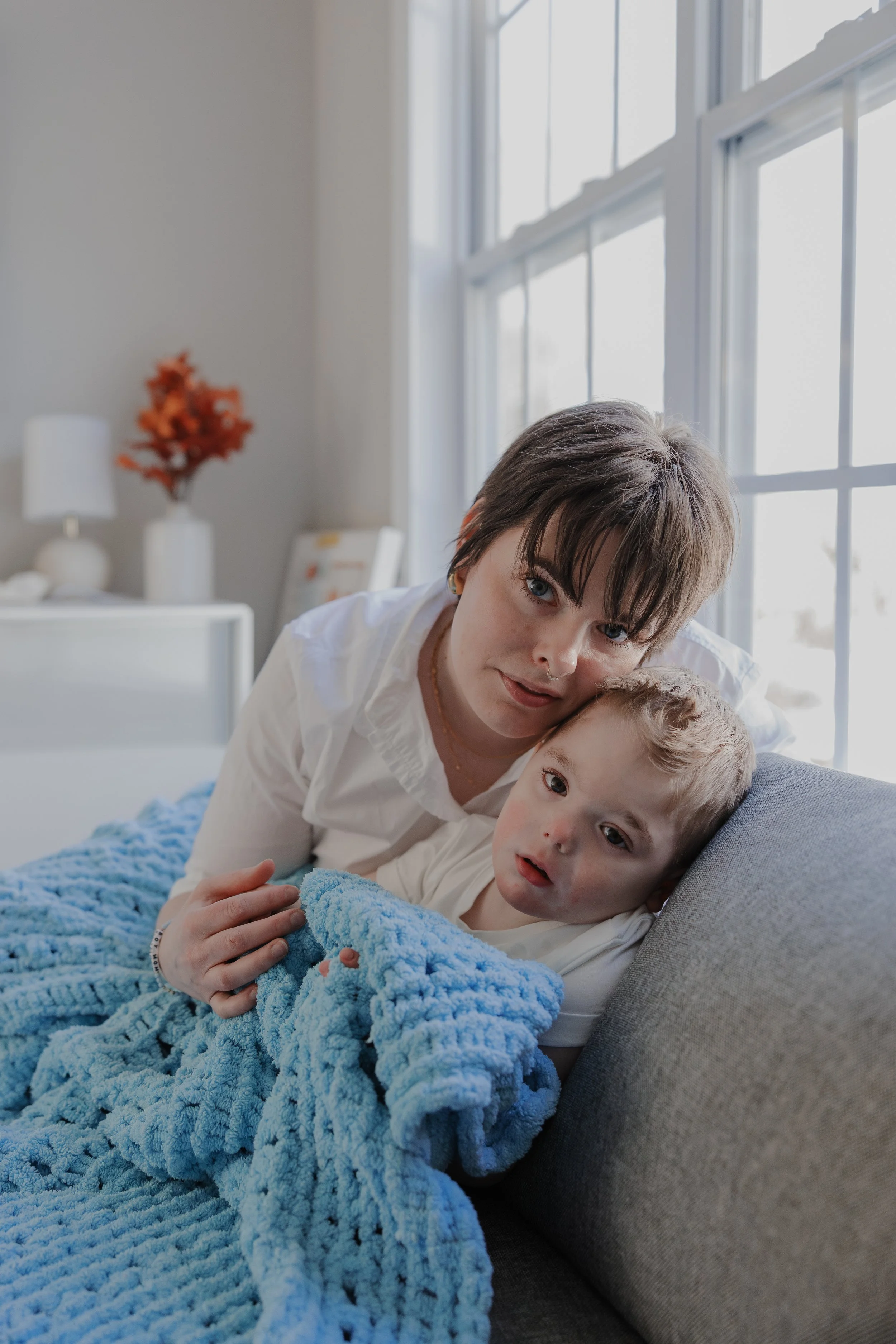 Mother cuddles her son wrapped in a soft blue blanket near a sunlit window during an inclusive in-home family photography session in Durham, NC.