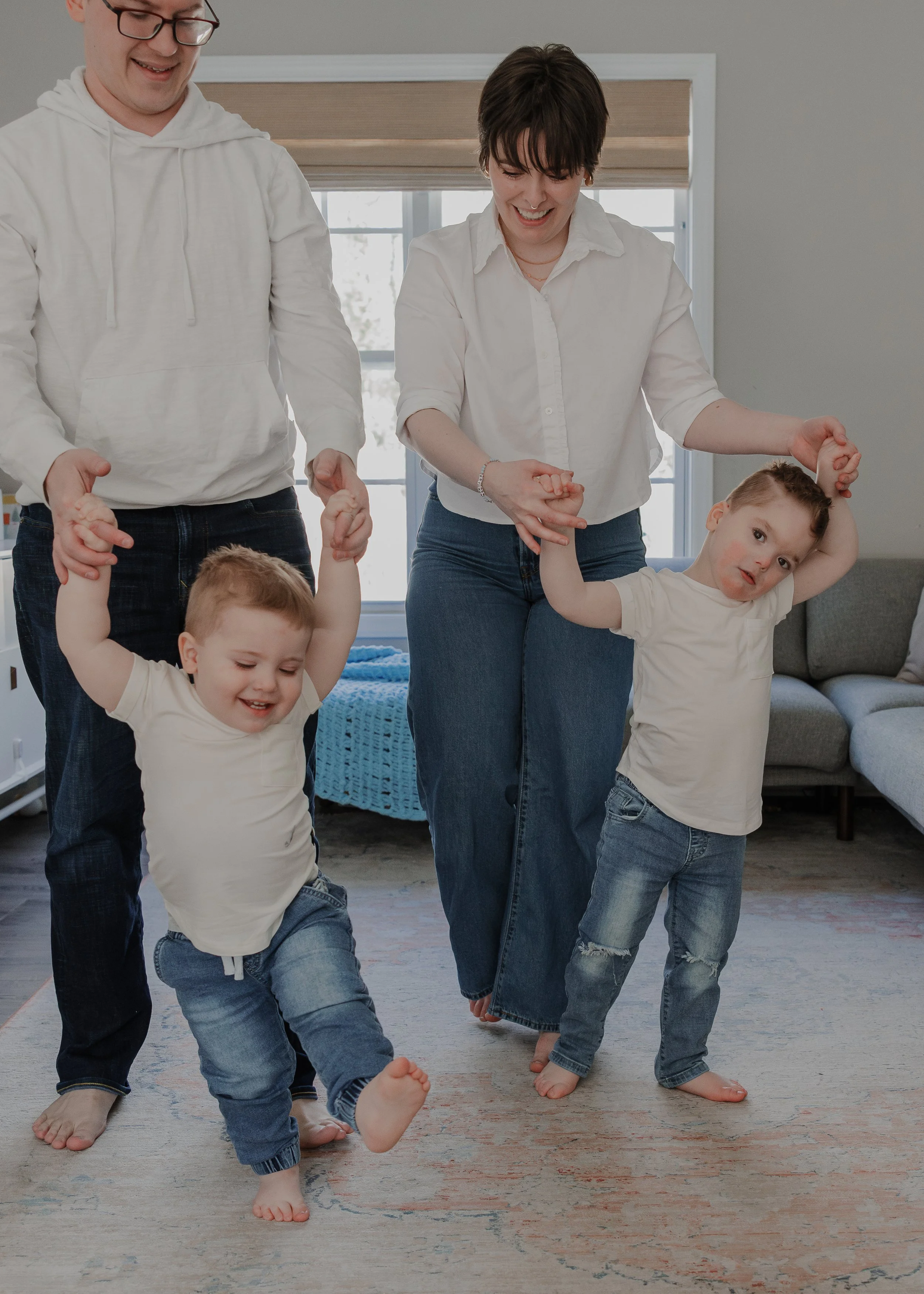 Parents hold their two young sons' hands while walking together across the living room during an inclusive family photography session in Durham, NC.