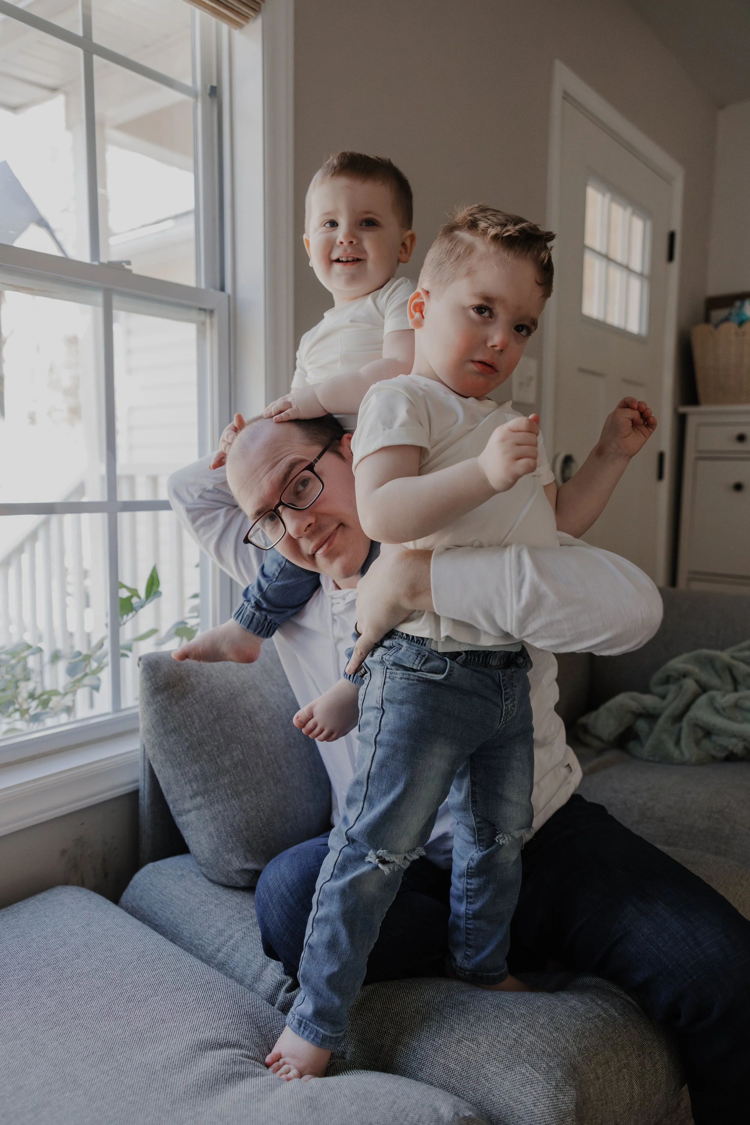 Father balances his two young sons on the couch beside a window during an in-home family photography session in Durham, NC.