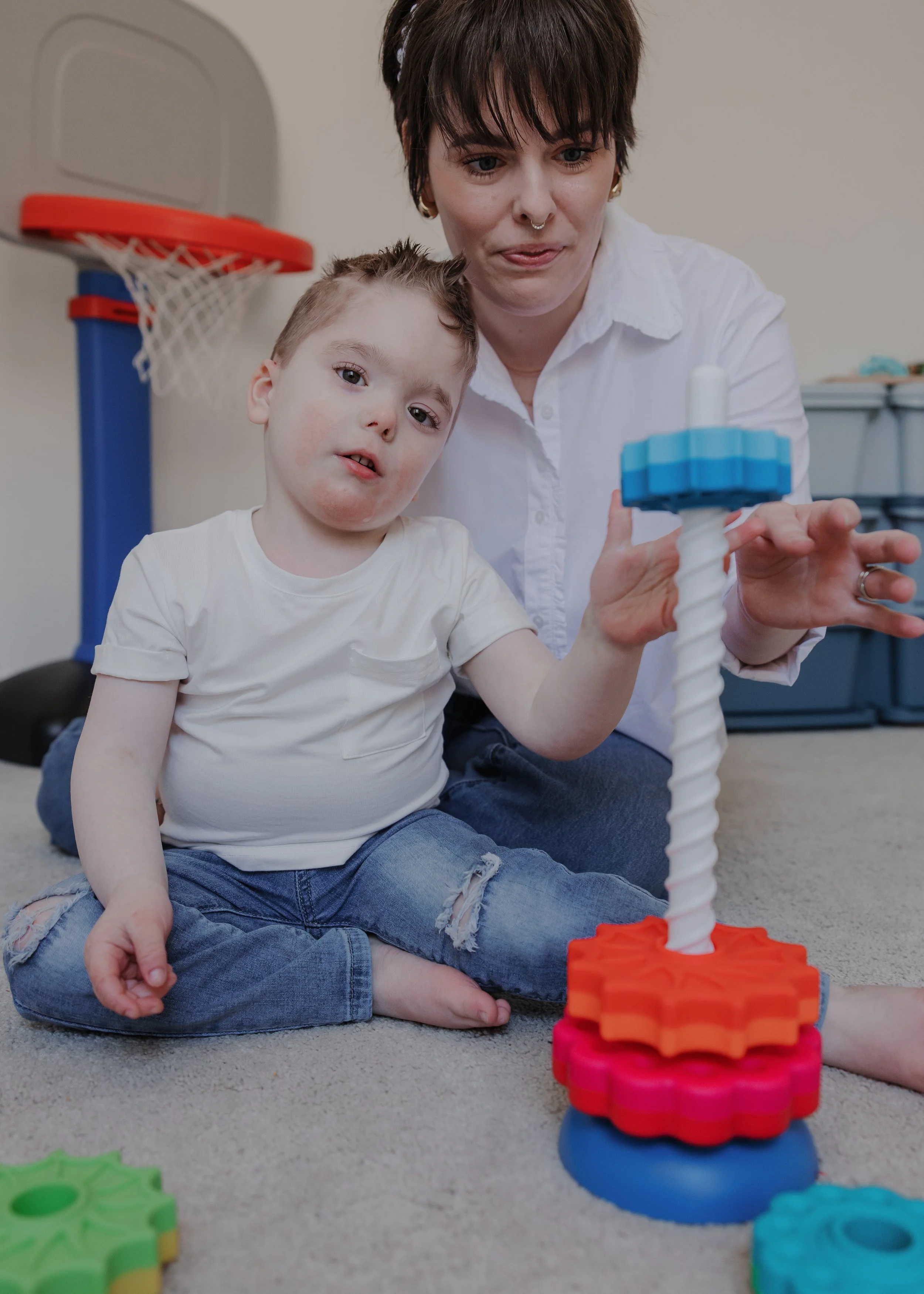 Mother sits with her young son as he plays with a spinning stacking toy during an inclusive in-home family photography session in Durham, NC for a child with ADNP Syndrome.