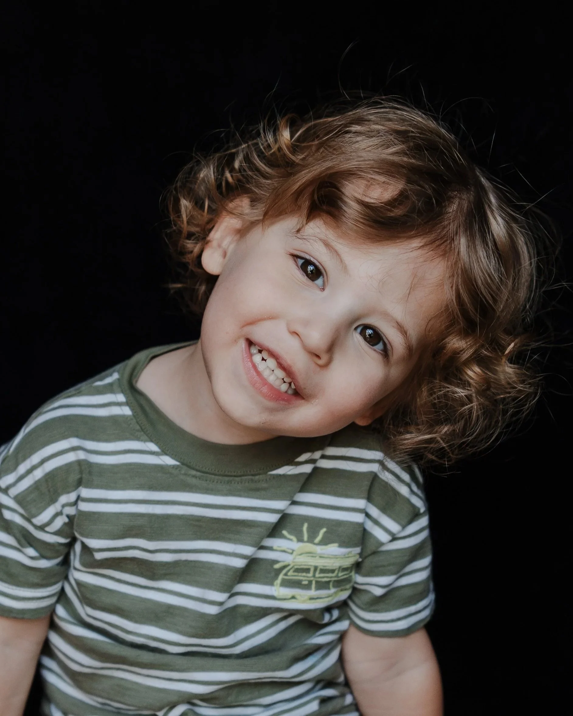 A young boy tilting his head and smiling against a timeless black background, captured by a Raleigh school photographer.