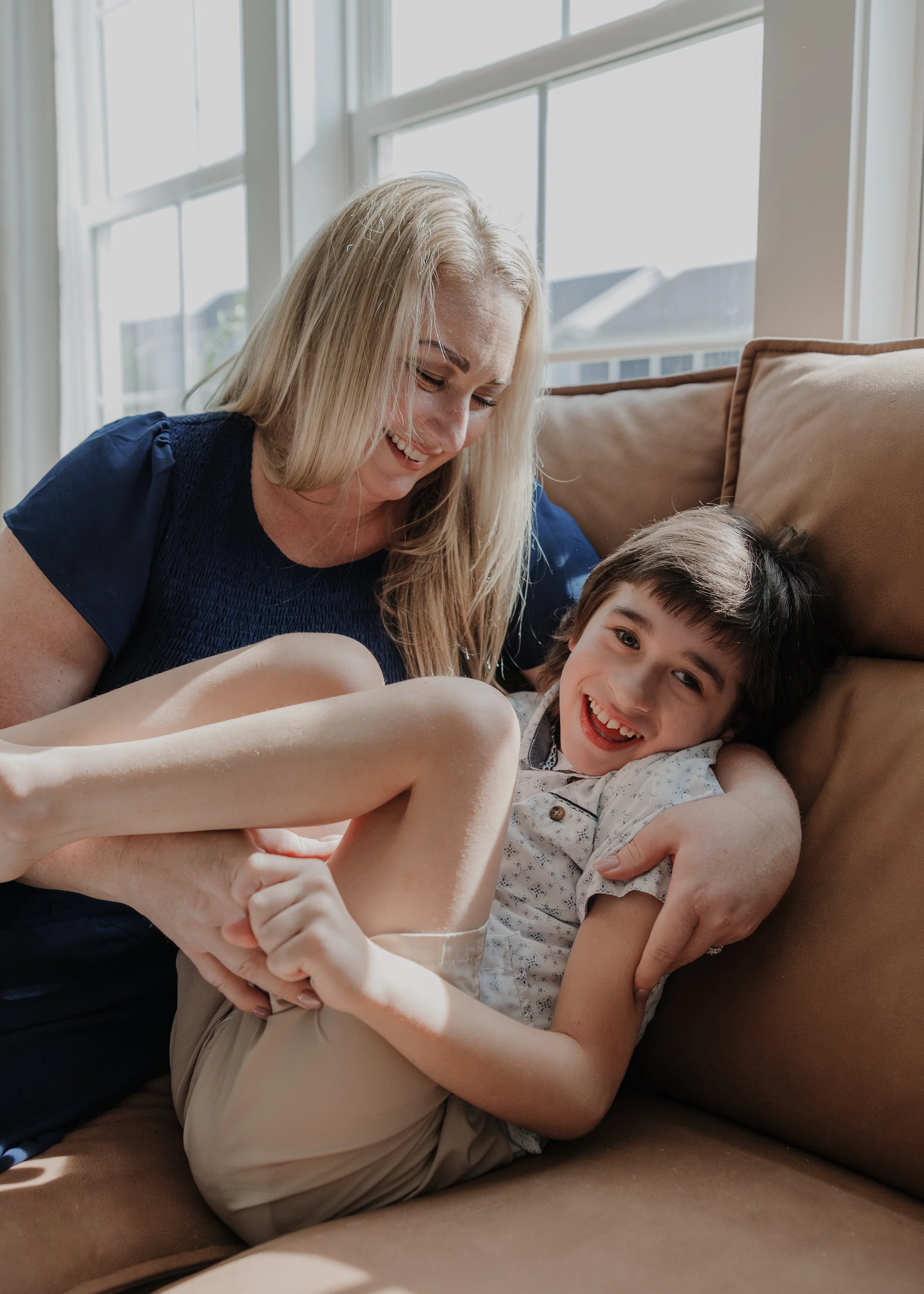 A mother laughing and cuddling with her son on a sofa, showcasing the joy of an inclusive family photo session.