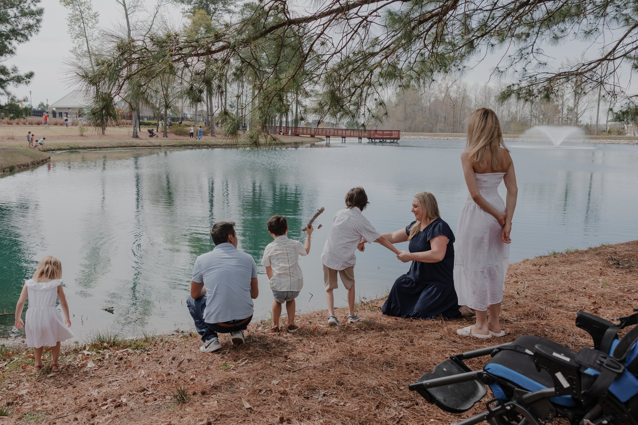 A cinematic outdoor shot of a family interacting by a pond in Knightdale, featuring their son and his medical equipment in the foreground.