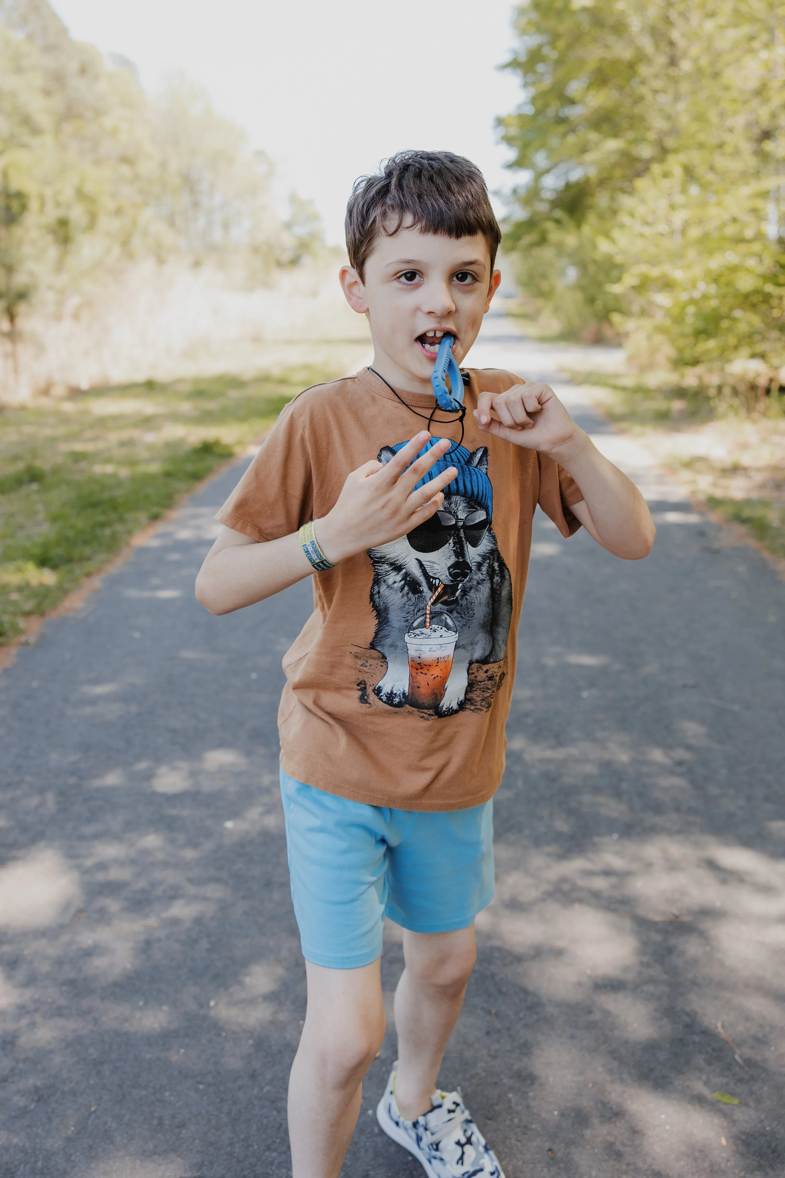 A profoundly autistic, largely nonverbal boy walks along a shaded Wake Forest greenway while chewing on a sensory tool during a child-led family photo session.