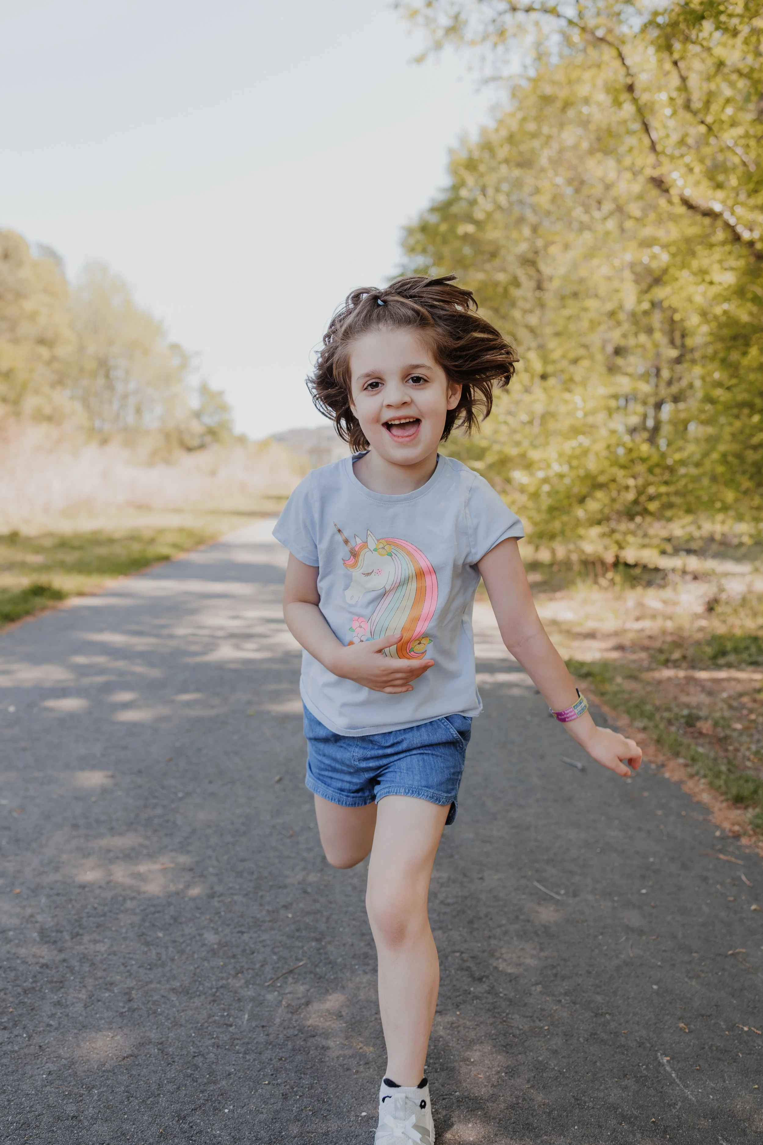 A young autistic girl with childhood apraxia of speech runs and smiles on a shaded Wake Forest greenway during a playful, child-led family photo session.